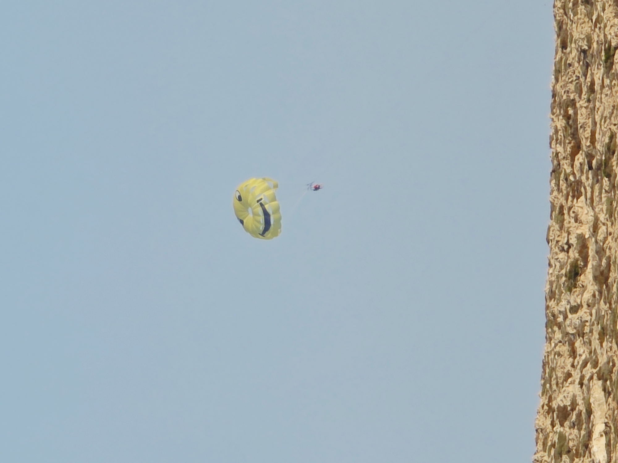 An object, identified as a smiley face parachute, is airborne against a clear light blue sky. A person is tethered to the parachute by a rope, appearing as a small red figure suspended below it. In the foreground, a textured landscape of light brown rocks with sparse green vegetation is visible. The scene is set in Ayia Napa, Cyprus, suggesting a coastal or cliffside location.