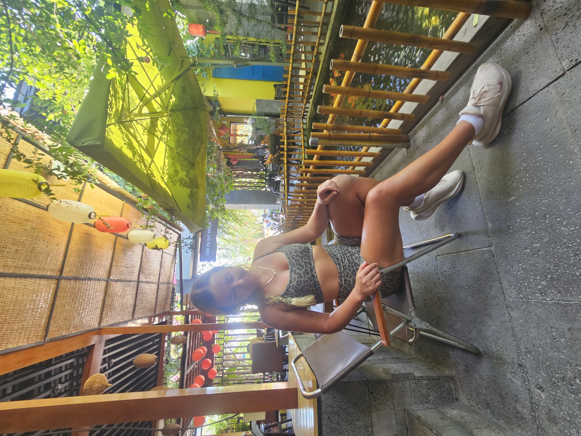 A young woman with blonde hair braided down her back, wearing a leopard print sports bra and matching shorts, sits on a folding metal and wood chair. She is in an outdoor seating area with a thatched roof supported by wooden beams. In the foreground, to her right, is a bamboo fence bordering a shallow body of water with koi fish visible. Behind her, the seating area continues with more tables and chairs, and a backdrop of lush greenery. Red lanterns are visible hanging from the ceiling and in the distance. The overall setting suggests a cafe or restaurant in Mũi Né, Vietnam, possibly near a body of water.