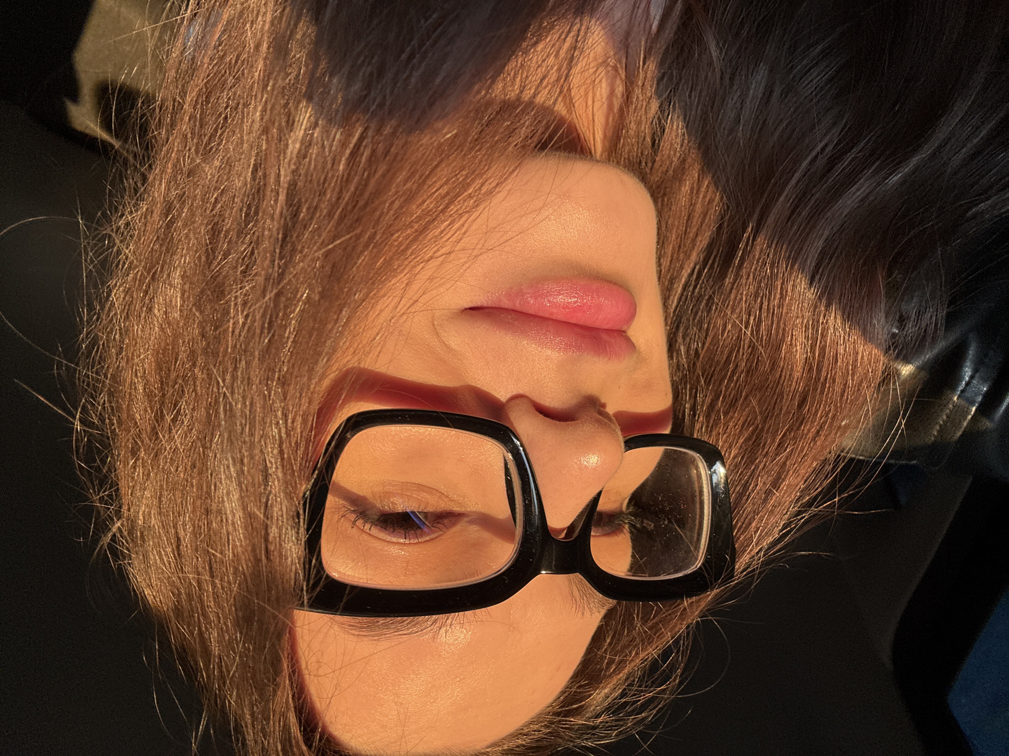 A close-up portrait shows a person's face, tilted and framed by long, light brown hair that flows around the head and across a dark, indistinct background. The subject is wearing thick, black-framed glasses. One eye is visible, appearing closed or looking downwards, while the other is mostly obscured by the glasses' bridge. The lips are slightly parted, showcasing a soft, natural pink color.
The scene is bathed in warm, golden sunlight, characteristic of the golden hour (late afternoon or early morning), which highlights the contours of the face, the texture of the skin, and individual strands of hair. This strong, directional light creates a glowing effect on the cheek, nose, and forehead. The overall mood is peaceful and serene, suggesting the person is resting or in a reflective state. There are no specific location cues visible within the image to identify the provided context of Izhevsk, Russia. No text is visible in the media file.