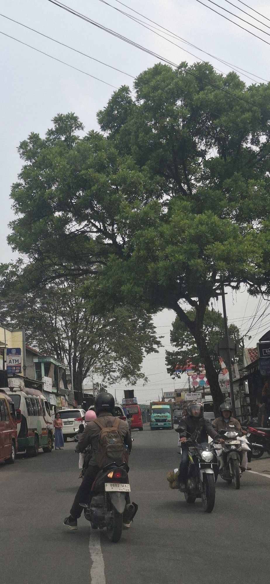 The image captures a street scene in Sirnagalih, Indonesia, under an overcast sky. The foreground features two motorcycles in motion, with riders wearing helmets. The motorcycle on the left is closer to the viewer, and its license plate reads "F 0002 WAZ 04-20". The rider is wearing a brown jacket and a backpack. A second motorcycle, with a rider also wearing a helmet, is to the right and slightly ahead, its headlight illuminated.
In the mid-ground, traffic is present, including a white car, a red van, and a turquoise truck with "TOKO BERAS D2" visible on its side. A person in a pink shirt and blue pants stands on the left side of the road. A large, leafy tree dominates the upper half of the image, its branches extending across the sky. Various buildings and shops line the street on both sides, with signs for "ATM" and other businesses visible. Electrical wires are strung across the top of the frame, and a utility pole is present on the right. The road is paved and has a white line marking. The overall impression is of a busy, ordinary day in a residential or commercial area.