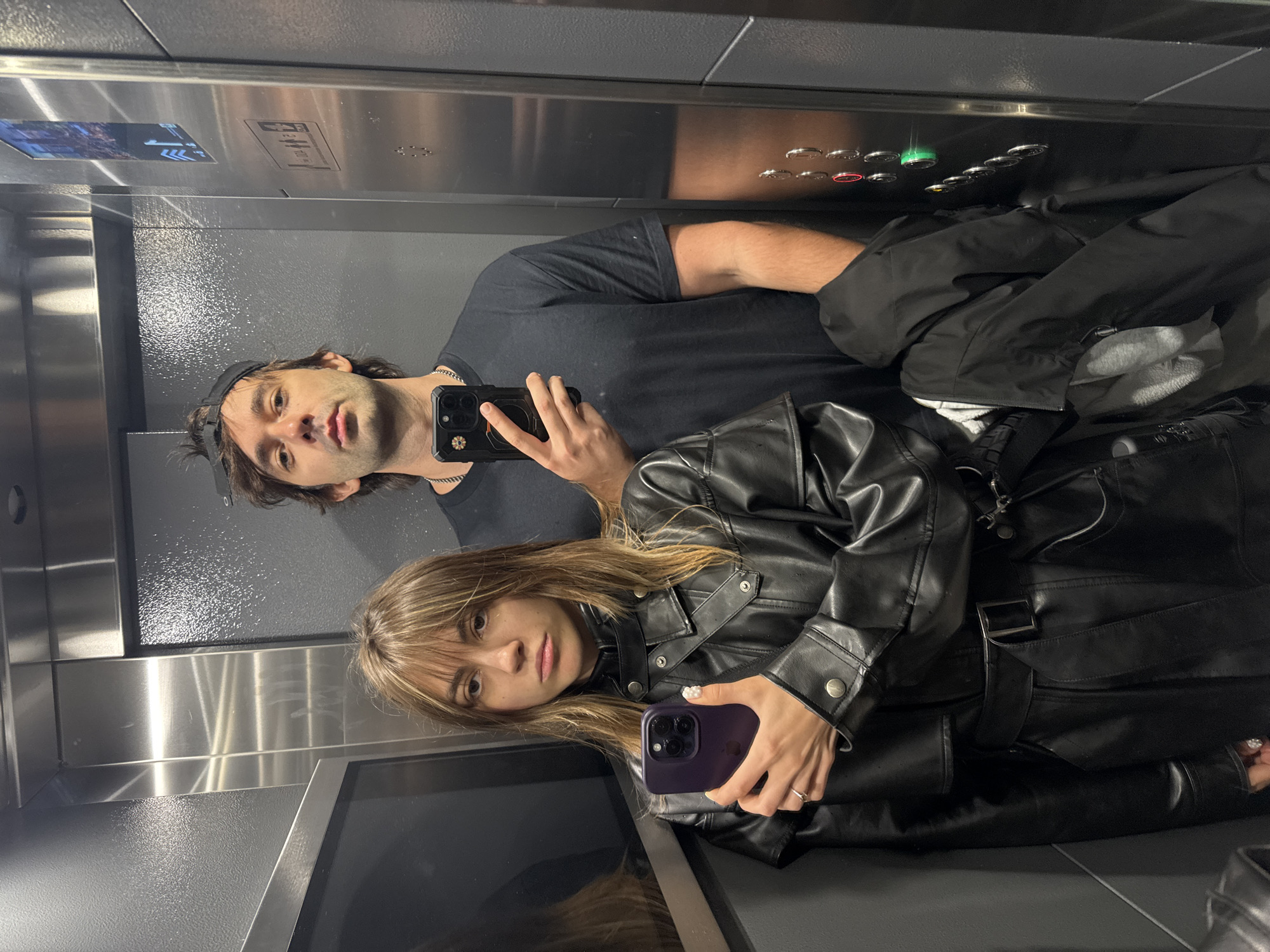 Two people, a man and a woman, are taking a selfie in an elevator. The woman is on the left, holding a purple smartphone and wearing a black leather trench coat. The man is on the right, holding a black smartphone and wearing a black t-shirt and a black baseball cap. The elevator has stainless steel walls and buttons on the right side. The image is a selfie taken from inside the elevator. The location is not specified in the image or its metadata.