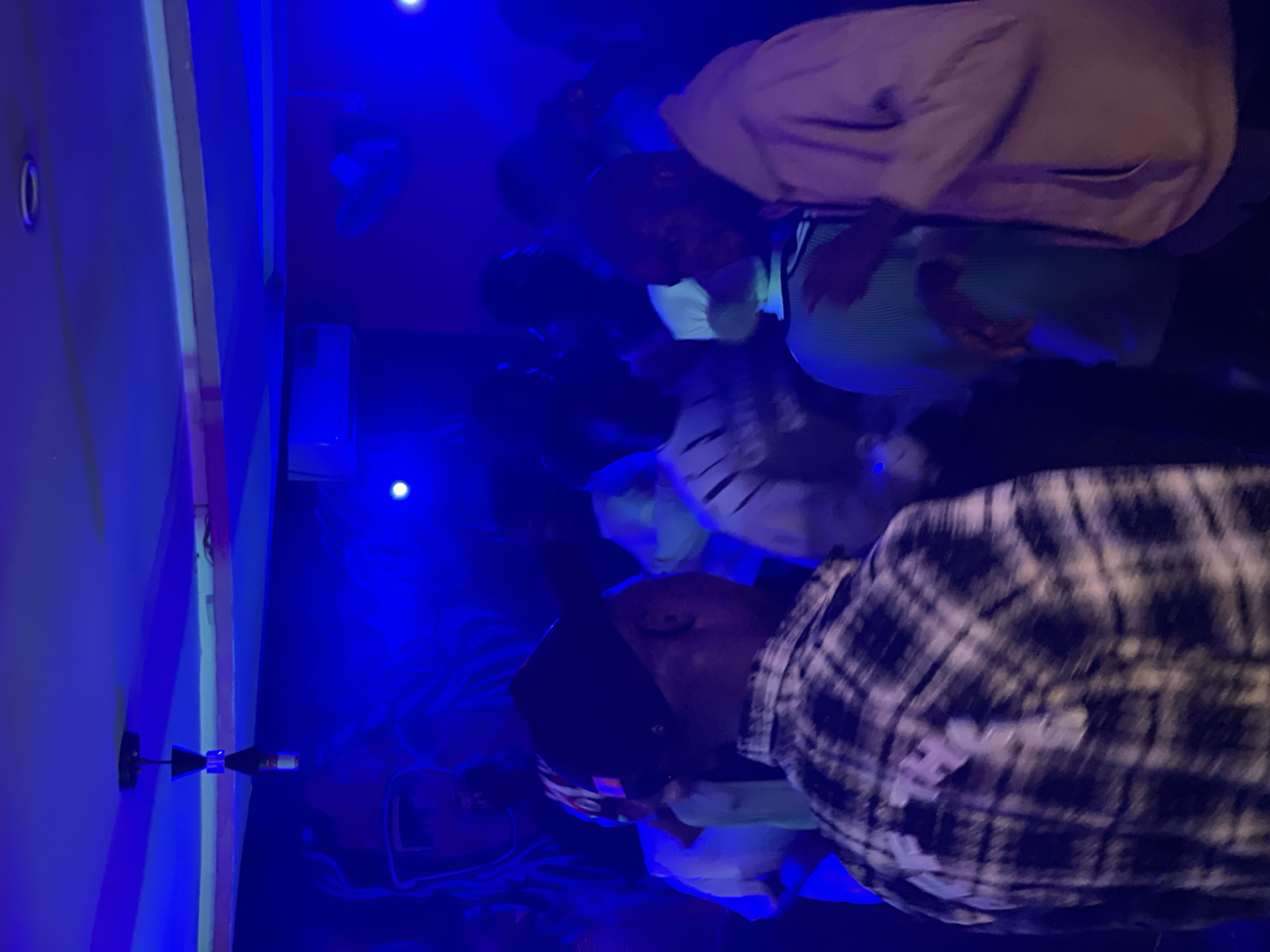The image depicts an indoor gathering illuminated by blue lighting, likely a club or party scene. In the foreground, a person wearing a black baseball cap and a black and white plaid shirt is seen from behind. To their right, another individual in a light-colored hoodie is visible from the side. Several other people are present in the background, their forms blurred by the low light and movement. The environment appears to be decorated with a mural on the wall, partially visible, and a hanging light fixture. An air conditioning unit is mounted on the wall. The overall atmosphere is dimly lit and energetic, characteristic of a nightlife venue in Durumi, Nigeria.
