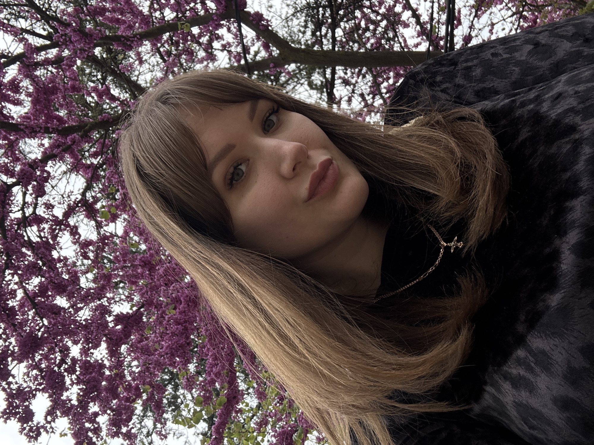 A young woman with long, light brown hair poses for a selfie outdoors. She is facing the camera at an angle, with her head tilted slightly. She has fair skin, green eyes, and full lips. She is wearing a dark-colored, patterned top and a delicate gold necklace with a small cross pendant. Behind her, a tree with vibrant purple flowers creates a colorful backdrop. The sky appears overcast. The location is Yevpatoriya, Ukraine.