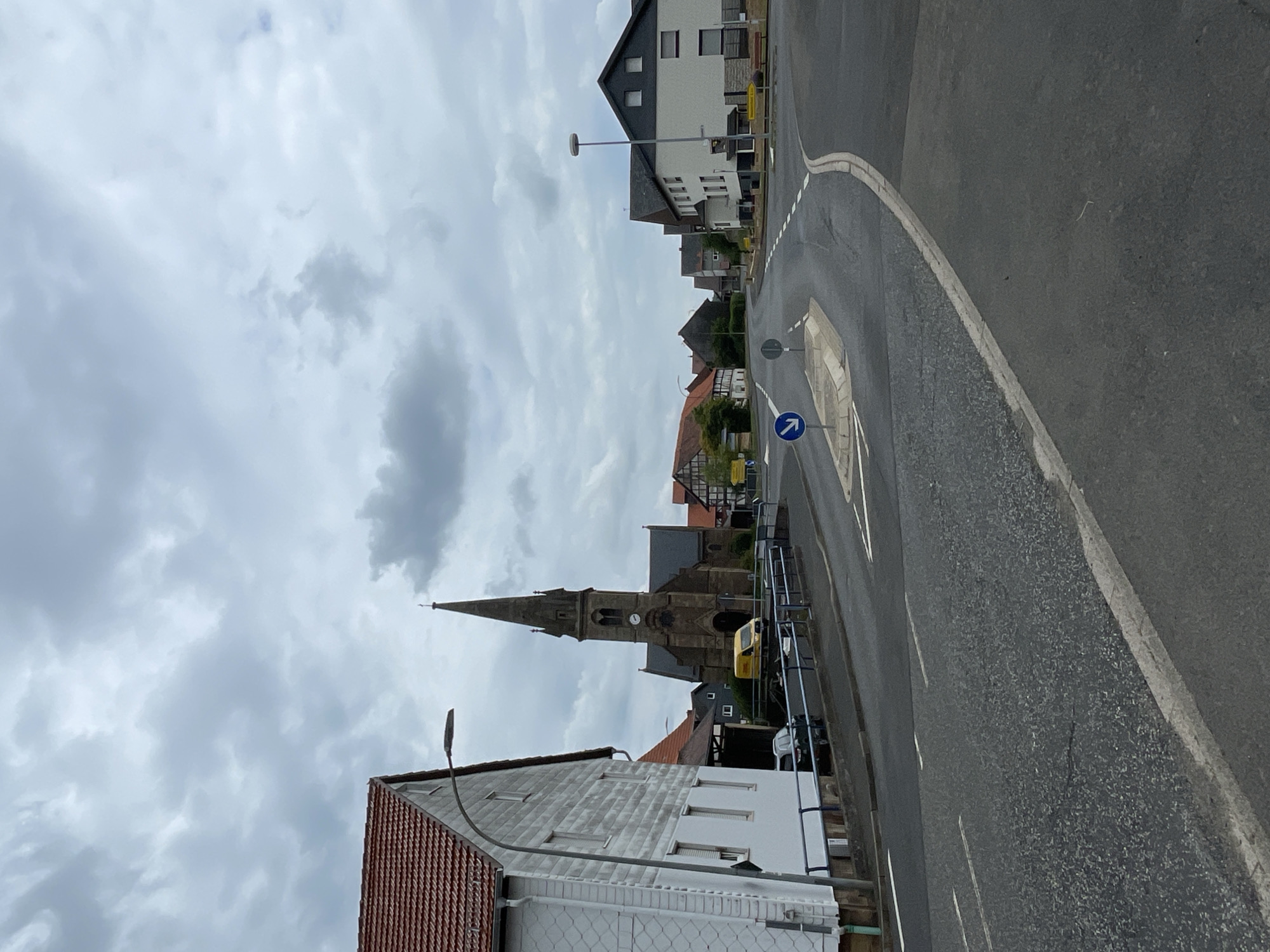 The image depicts a street scene in Seigertshausen, Germany, under a cloudy sky. The photograph is oriented vertically. In the foreground, a white building with a red-tiled roof and a metal fence is visible. To the left of this building, a street lamp stands. Behind this structure, a tall, pointed church spire dominates the middle ground, with a clock face visible on its tower. The church is made of stone and has arched doorways and windows. A yellow van is parked near the church. Further back, several houses and buildings line the street, showcasing a mix of architectural styles, some with modern dark roofs and windows, and others with traditional red tiles. The street itself is paved and curves to the right. A blue traffic sign with a white arrow indicates a mandatory right turn. The overall impression is a quiet, residential area, likely in the afternoon, given the overcast conditions. There is no visible text in the image, other than on the traffic sign.