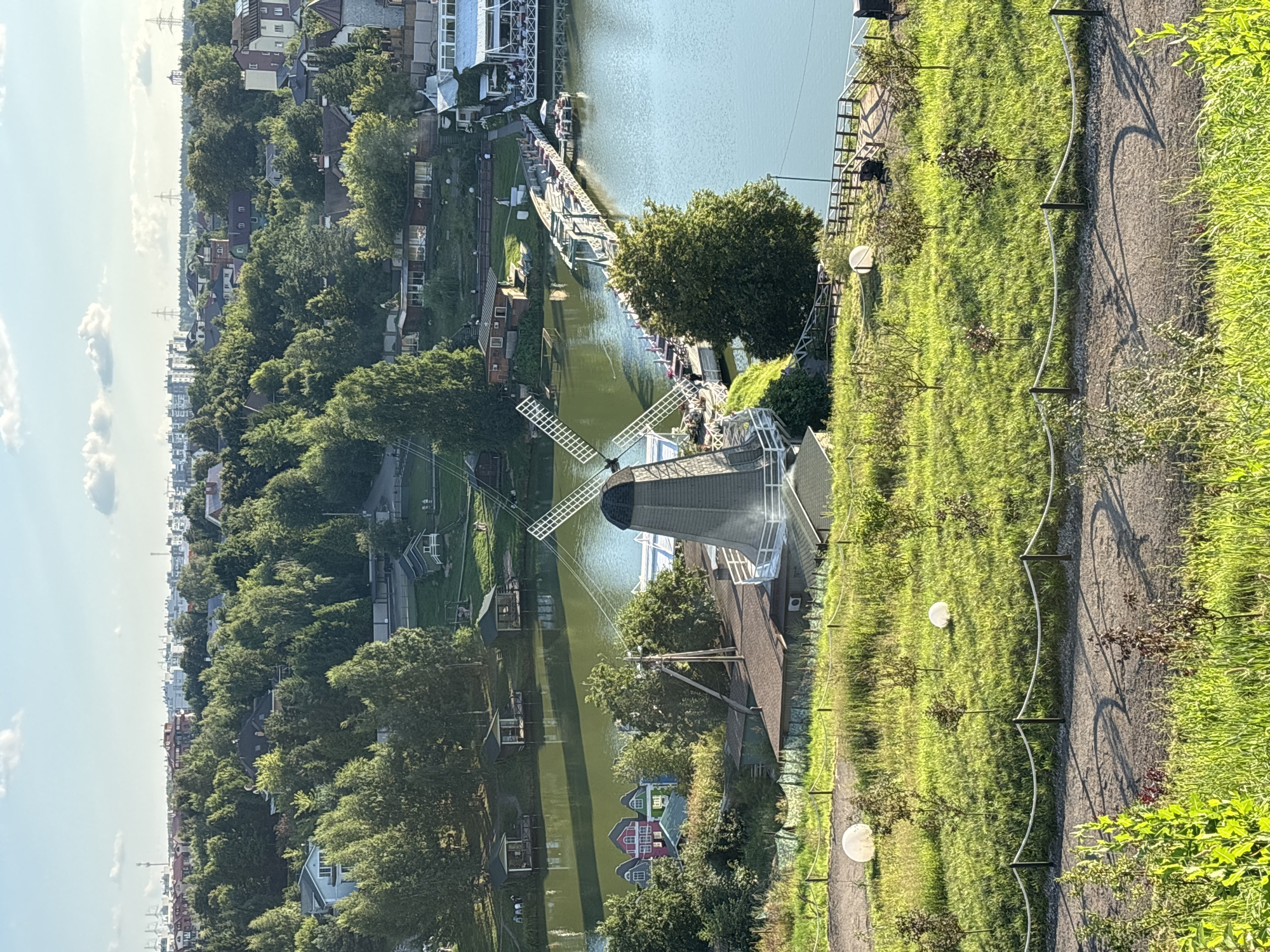 The image depicts a scenic view of a park or recreational area, likely located in Romashkovo, Russia, with a body of water as its central feature. A prominent Dutch-style windmill stands on the shore of the water, its blades partially visible. Adjacent to the windmill is a wooden bridge that extends across the water, connecting to buildings on the opposite bank. Several small, colorful houses are clustered on the left side of the water, near the windmill.
The foreground of the image is a grassy slope dotted with young trees and illuminated by two large, spherical white lights. A gravel path runs along the bottom of the slope, bordered by a rope fence. In the middle ground, the calm, greenish water reflects the surrounding trees and structures. The background reveals a dense forest bordering the water, with a cityscape of buildings and power lines visible in the distance under a partly cloudy sky. The overall impression is one of a peaceful, well-maintained outdoor space, possibly a tourist attraction or a leisure facility. No specific human actions or interactions are clearly discernible.