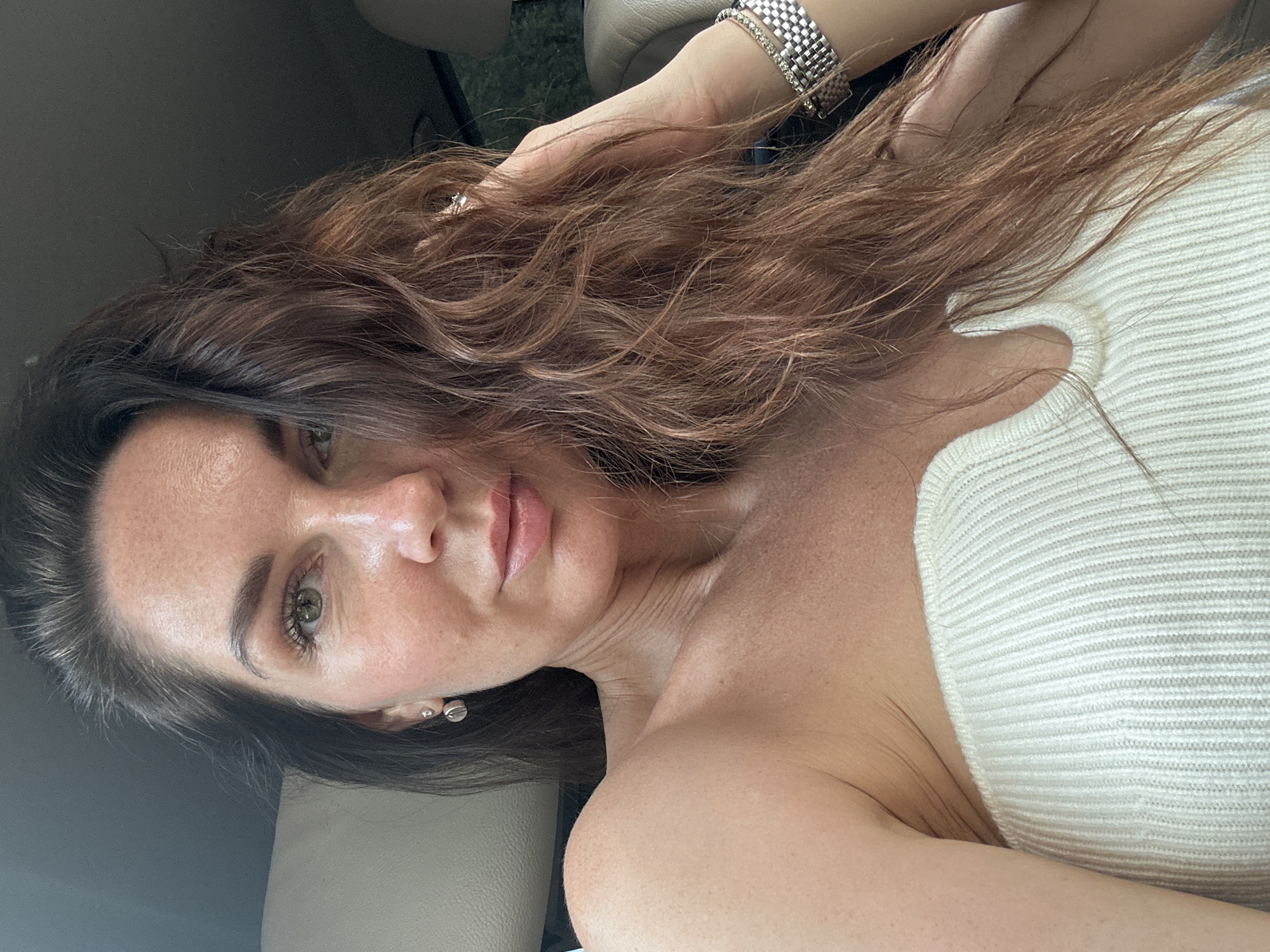 The image shows a woman with brown hair, wearing a white strapless top. She has fair skin with freckles, green eyes, and is wearing earrings and a bracelet. Her hair is partially covering her face. The background appears to be the interior of a car, with light-colored seats visible. There is no indication of the location being Chaikovsky, Russia.