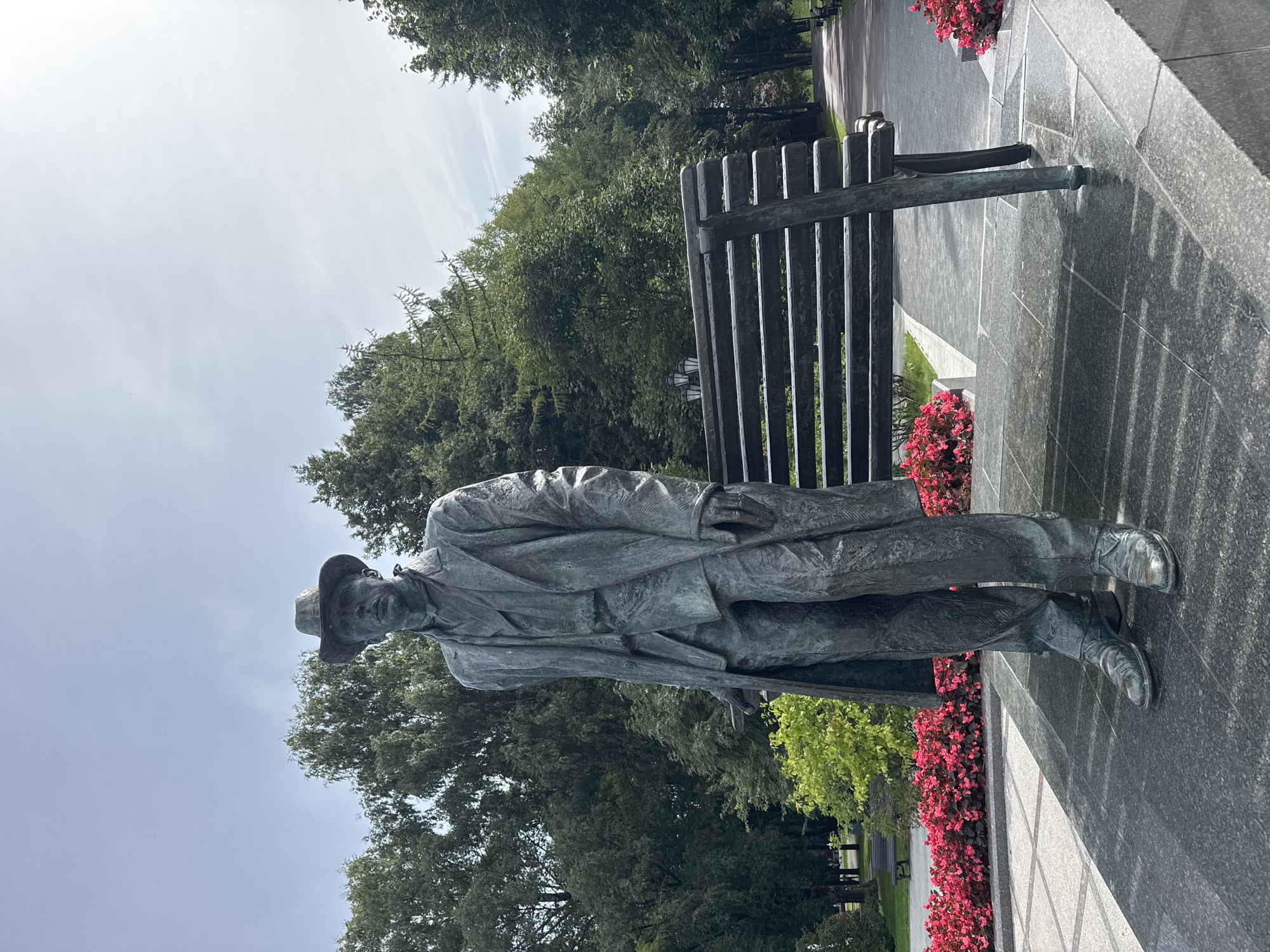 The image shows a full-body bronze statue of a man wearing a hat, a long coat over a suit, and shoes. He is standing next to a park bench. In the background, there are trees and a path. The statue is located in a park setting. The statue is positioned in the foreground on a dark granite base. The man appears to be standing at ease, with his left hand resting near his side and his right hand holding a thin object, possibly a cigarette or a cane. The bench behind him is made of dark metal with a slatted seat and back. There are beds of red flowers on either side of the statue and the bench. The background consists of lush green trees, some of which appear to be coniferous. A paved pathway is visible in the distance, leading through the park, with additional benches and trees lining the sides. The sky above is bright with some wispy clouds. This statue is located in the Alexander Garden in Veliky Novgorod, Russia, and depicts the poet Georgy Dedyushko.
