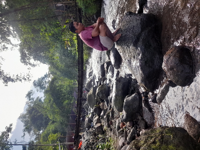 A man in a pink shirt and light shorts sits cross-legged on a large, dark rock in a shallow river. He faces away from the camera, looking down the river. The water flows around and between many smaller rocks. Lush green trees line the riverbanks. A wooden bridge, with people standing on it, spans the river in the background. Sunlight streams through the trees.