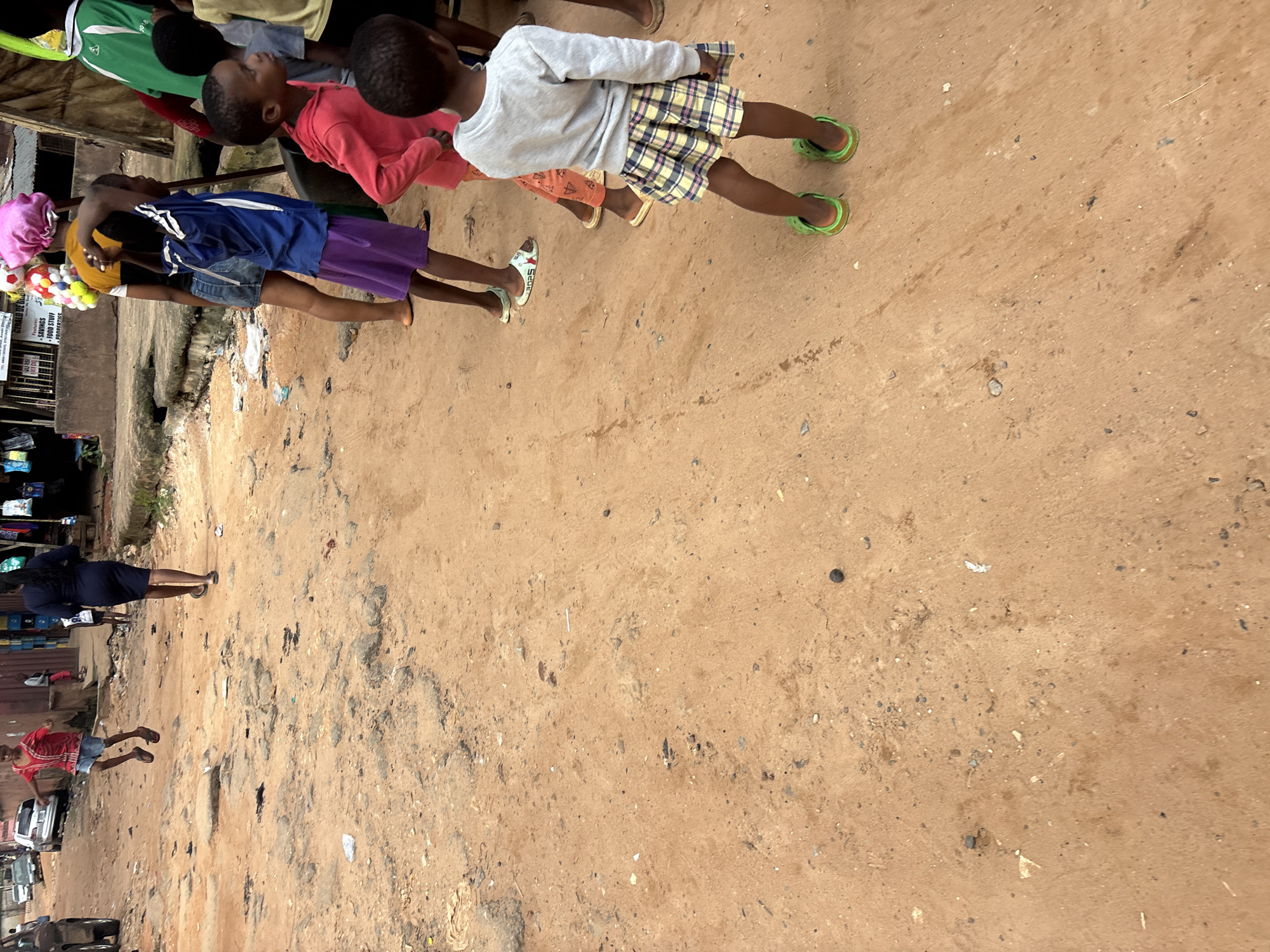 The image depicts a scene on a dirt road in Onitsha, Nigeria, likely in a market or residential area, characterized by uneven terrain and scattered debris. Several individuals, predominantly children, are present. In the foreground, a child wearing a plaid skirt and green clogs is facing away from the camera. To the left, another child in a pink top and orange pants is visible. Further back, a child in a blue shirt and purple skirt is captured in motion.
Several other children are in the mid-ground, including one in a red shirt and shorts who appears to be mid-jump or running, and another in a blue and white striped shirt. A woman in a dark dress is walking away from the camera towards the background. In the distance, a few cars are parked on the side of the road. The environment includes buildings with corrugated metal roofs and stalls displaying various goods, suggesting a commercial setting. The overall impression is of a dynamic street scene with active participation from children.