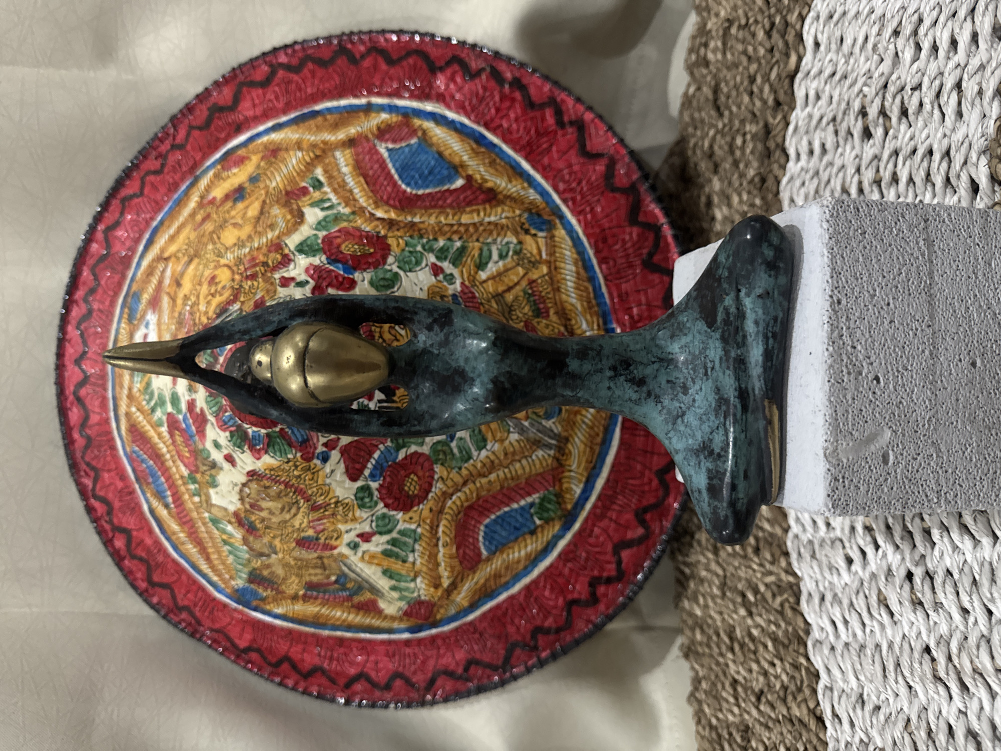 A high-angle shot shows a bronze sculpture of a bird, its beak pointing left. The bird cradles a golden egg in its body. The sculpture is positioned on a decorative, circular plate with an ornate design, including images of figures and floral motifs. The plate has a red and black patterned rim. The sculpture's base rests on a rectangular gray stone, next to a woven mat featuring both light and dark brown strands. The backdrop includes a cream-colored fabric and a wall. This image was taken in the Ubud District of Indonesia.