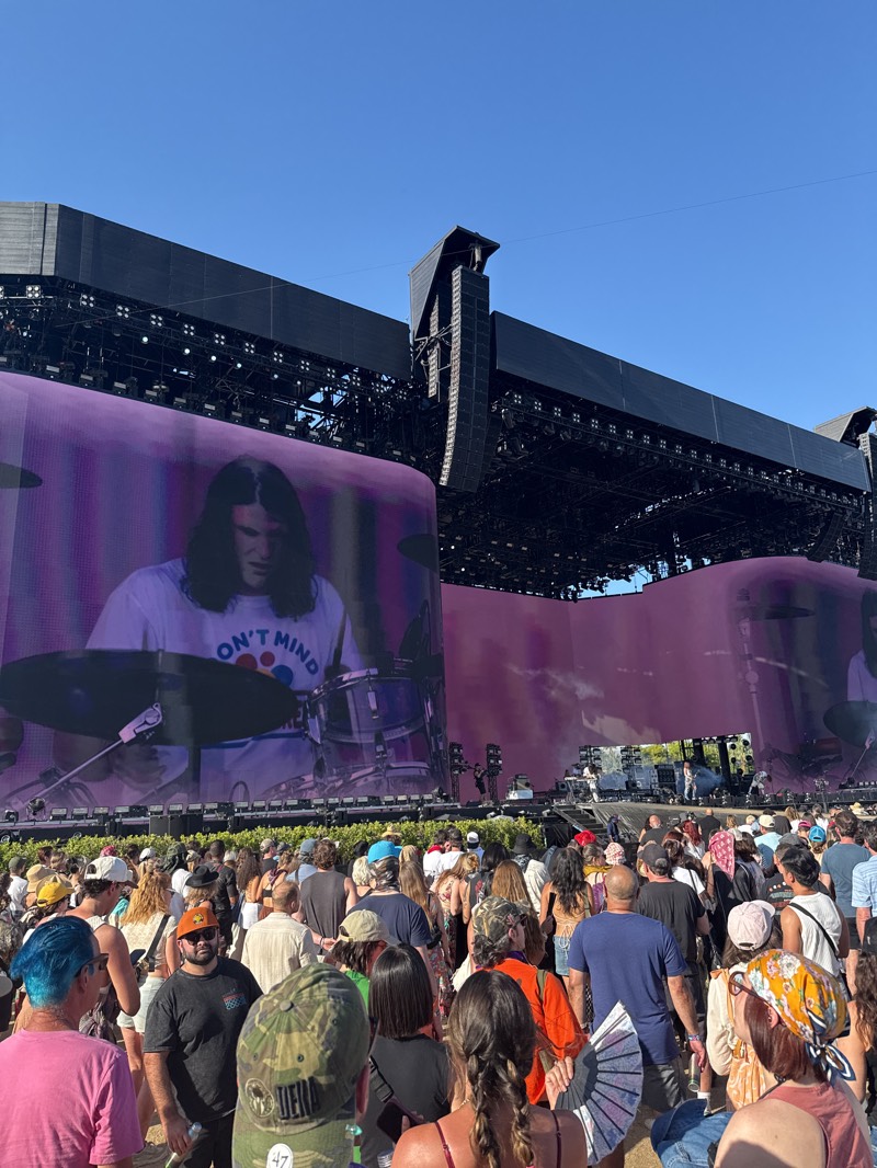 The image depicts a large crowd of people gathered at what appears to be an outdoor music festival in Indio, California, United States. In the background is a large stage with massive video screens showing a drummer. The stage has large speaker arrays and overhead structures. The crowd is diverse, with people of various ages and ethnicities. Most people are standing and facing the stage. The sky is clear and blue, suggesting a sunny day. The overall atmosphere seems to be lively and engaging, consistent with a music festival environment.