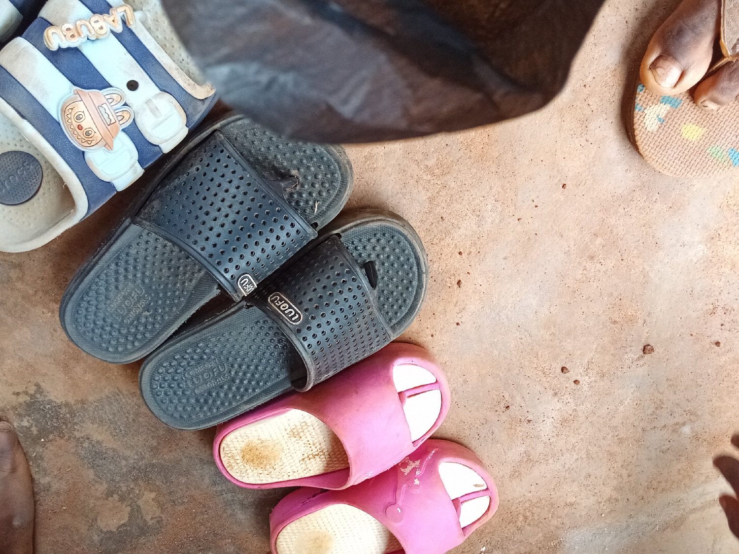 Multiple pairs of casual footwear are scattered on a light brown, dusty concrete or cement ground. The scene is viewed from an overhead perspective.
In the upper left, a pair of blue and white striped slides is visible. They feature a cartoon character, possibly a bunny or bear, on the strap, and the word "LABUBU" is written on the inner side of the strap. A circular white detail on the left slide also shows the word "HOSS".
Below these, a pair of dark grey or black slides is positioned, one slightly overlapping the other. These slides have perforated straps and textured footbeds. The words "LUOFU ORIGINALS" are visible on the footbed of one slide, and "LUOFU" is printed on the strap of both.
Towards the bottom right, two pink slides or slippers are partially visible. Their interiors appear somewhat worn or stained.
Parts of human feet are also visible in the image. On the far right, toes wearing a patterned flip-flop (light brown with small colorful squares) are seen. A partial foot is also visible on the lower left corner.
A dark, possibly black, object, resembling a bag or cloth, casts a shadow over the upper central part of the frame. The ground itself is uneven and appears to have small grains or debris.
The image depicts a casual, everyday setting, likely outdoors or in a simple entryway, consistent with a domestic environment in Shagamu, Nigeria. No specific activity is taking place beyond the placement of the shoes. The lighting suggests daytime.