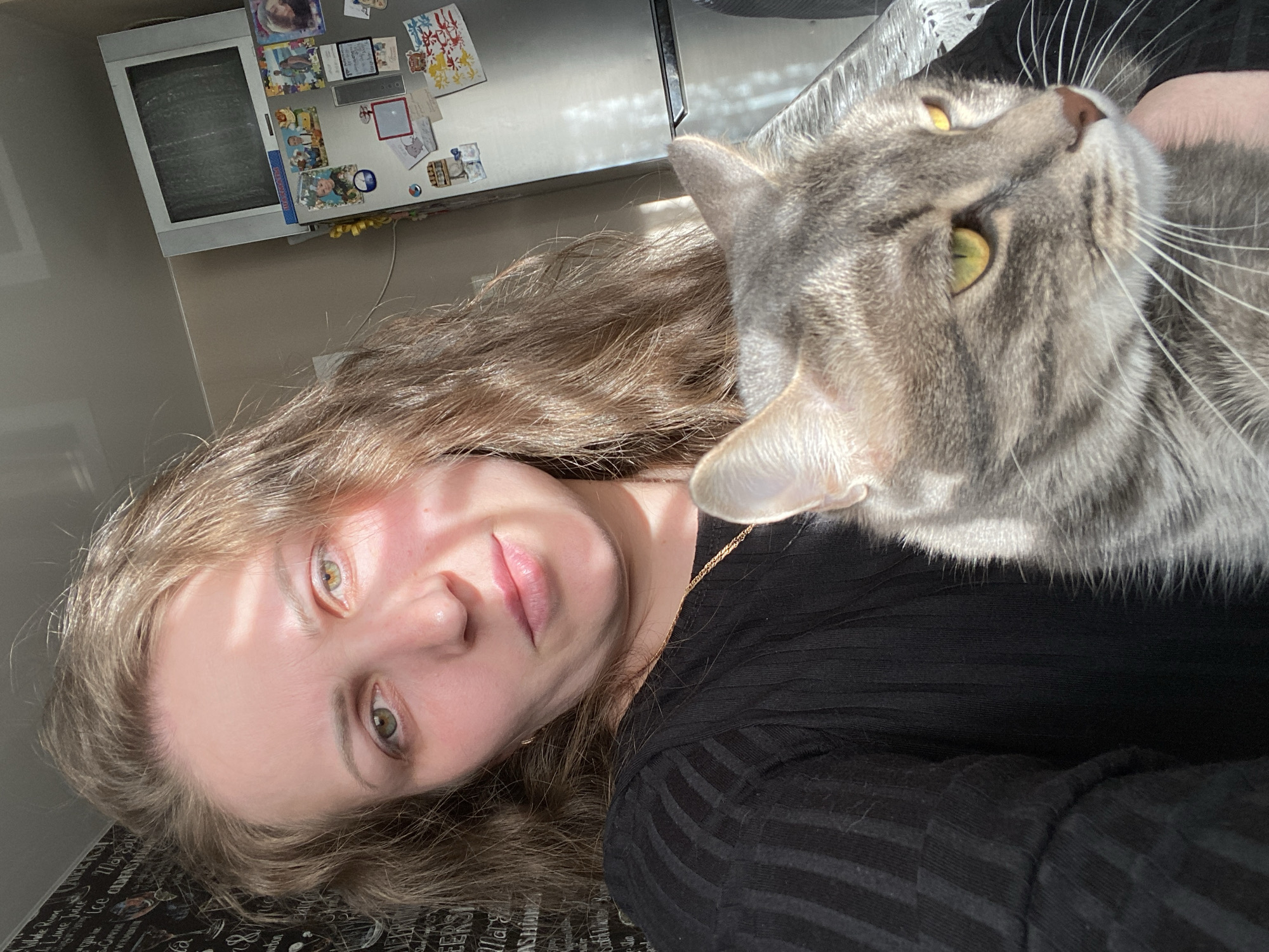 A woman with long, light brown hair is taking a selfie with a gray tabby cat. The woman has fair skin, green eyes, and is wearing a black shirt. The cat is looking upwards with yellow-green eyes. The background includes a refrigerator with pictures and magnets, a wall, and a surface with writing on it. Location cannot be determined.