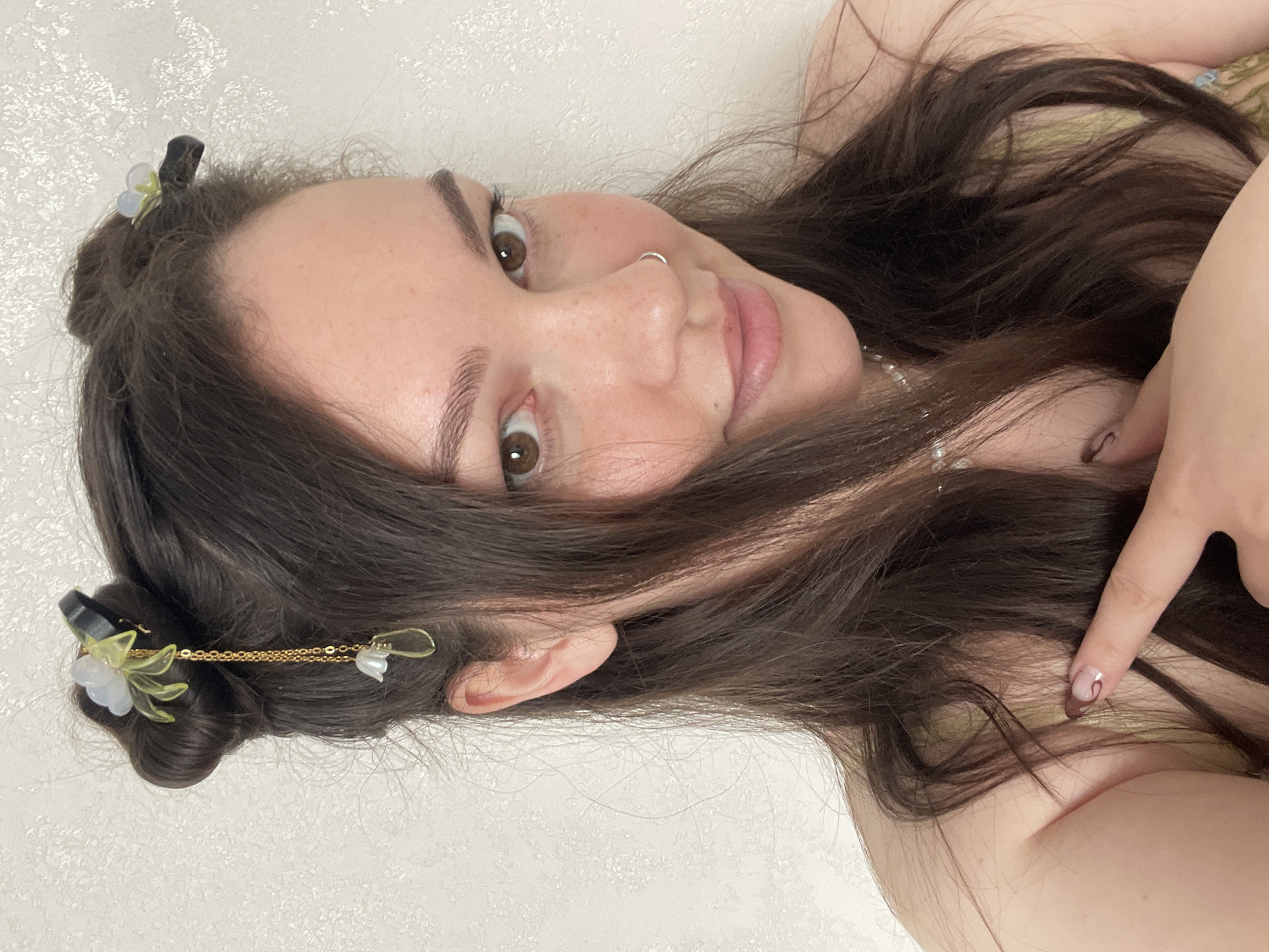 A selfie captures a young woman from a low-angle perspective, rotated as if she is lying down and looking up at the camera. She has long, dark brown hair, dark brown eyes, and visible freckles across her nose and cheeks. Her eyebrows are well-defined, and she has a small silver hoop piercing in her left nostril. Her lips are slightly parted in a soft, gentle smile, and her expression is engaging and content.
Her hair is styled with two small buns on top of her head, adorned with decorative hair clips. One clip features a white flower with translucent green leaves, and another, connected by a delicate gold chain, has a smaller white pearl or flower detail. The rest of her hair flows loosely around her face and over her shoulders. She appears to be wearing a light-colored, possibly pale green or yellow, garment. A string of pearls is visible around her neck, suggesting a necklace. Her right index finger is visible, resting near her shoulder, showcasing a fingernail with a light base and a distinct dark brown or reddish swirly design.
The background is a plain, light-colored wall with a subtle texture, suggesting an indoor setting. There are no other objects or distinguishing features in the background. The lighting is soft and even, highlighting her features without harsh shadows. While the provided context indicates Izhevsk, Russia, no specific visual cues within the image confirm this location. No visible text is present in the image.