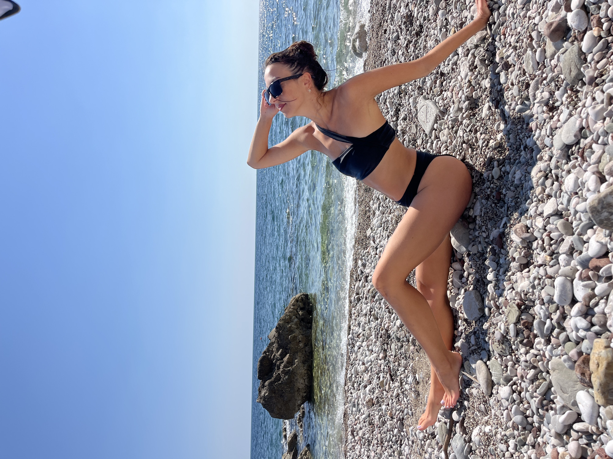This media file shows a woman in a black bikini on a pebbled beach in Bar, Montenegro. She is standing by the water, with her hand shielding her eyes from the sun. The water is clear and blue, with small waves gently lapping at the shore. The beach is covered in smooth, grey pebbles, and there are a few larger rocks scattered along the waterline. The sky is a clear, bright blue, suggesting a sunny day. The woman appears relaxed and enjoying the outdoors. The time of day appears to be daytime, given the bright sunlight and shadows. There is no visible text in the media.