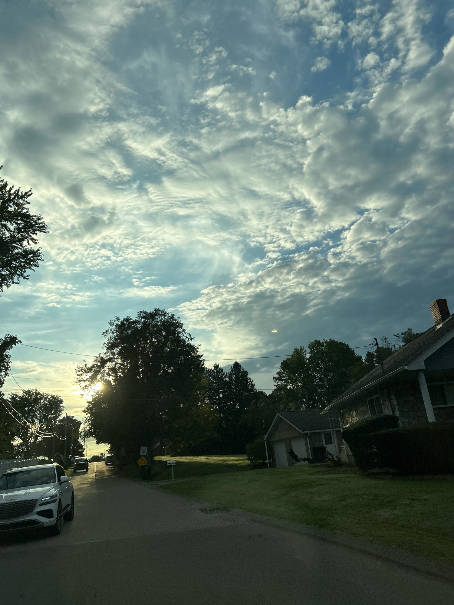 This is a roadside view in Robinson Township, United States of America, captured from a vehicle. The scene is dominated by a vast, cloudy sky with patches of blue and sunlight breaking through. Tall trees line the road, some silhouetted against the bright sky. In the foreground, a white SUV is parked on the left side of the asphalt road. Further down the road, other vehicles are visible. On the right side of the road, a house with a stone facade and a brick chimney is partially visible, with a manicured lawn in front. A detached garage is also present. The overall atmosphere suggests an outdoor setting, possibly during late afternoon or early morning due to the lighting.