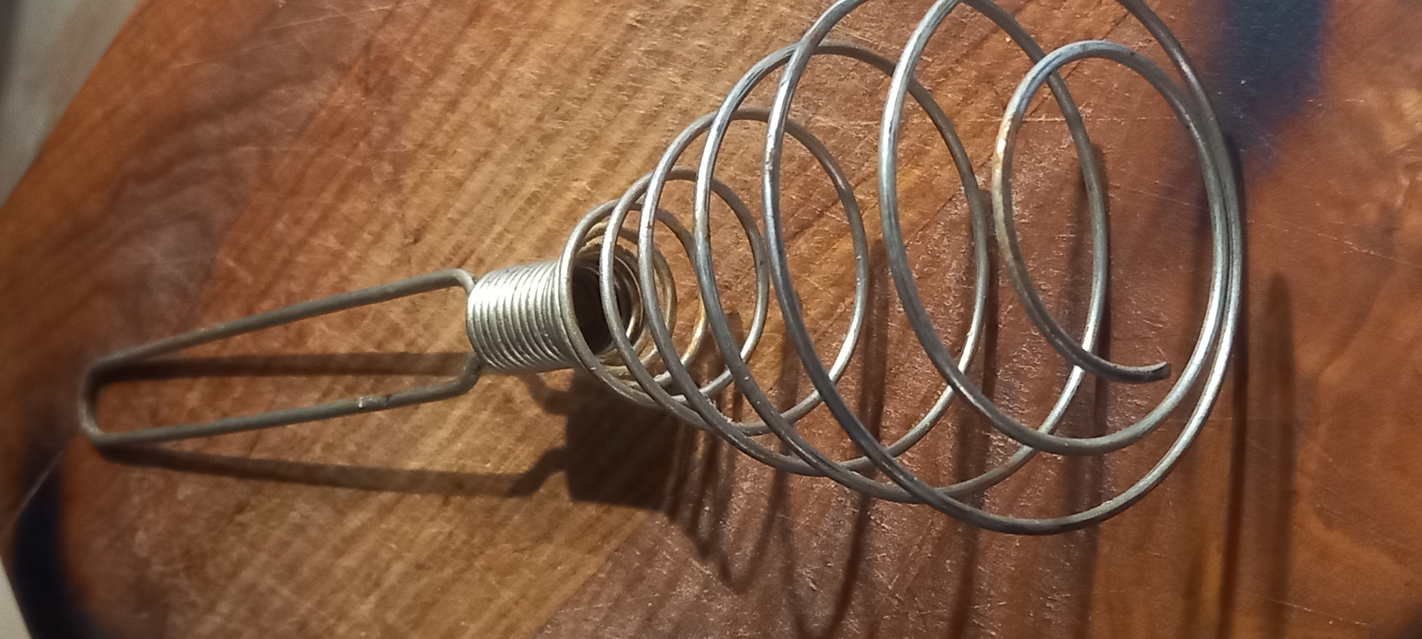 A silver-colored metal whisk is positioned on a worn brown wooden surface. The whisk is constructed from a continuous wire, forming a U-shaped handle, a tightly coiled spring midsection, and a broad, flattened spiral at its base. Sharp, elongated shadows of the whisk are cast to the right, indicating a light source from the left. The wooden surface displays visible grain patterns and minor surface abrasions. No individuals, actions, or interactions are depicted. This scene is identified as being in the city of Kryvyi Rih, Ukraine.