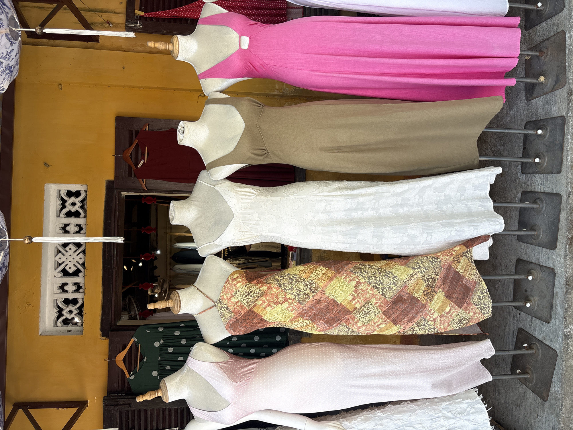 The image shows a storefront in Hoi An, Vietnam, displaying several dresses on mannequins. The storefront has a bright yellow facade with dark wooden window frames and shutters. Above the display, a decorative white carved screen is visible.
In the foreground, six mannequins are arranged in a row, each wearing a different dress. From left to right:
1. A pale pink, sleeveless, V-neck dress with a subtle polka dot pattern.
2. A halter-neck dress with a patchwork-style pattern in shades of brown, orange, yellow, and cream.
3. A sleeveless, white, textured lace or embroidered dress.
4. A sleeveless, taupe or light brown, simple slip-style dress with a V-neck.
5. A bright pink, sleeveless, V-neck dress with a cutout detail at the neckline.
6. A white dress is partially visible on the far right.
Behind the mannequins, the interior of the shop is visible, with racks of clothing and more dresses on display. Some of the clothing hanging inside appears to be red, green, and darker colored garments. Above the storefront, a blue and white patterned fabric or umbrella is partially visible, and white tassels hang down. The ground in front of the storefront is paved with grey stones. The overall impression is a colorful and inviting display of clothing in a traditional Vietnamese setting.