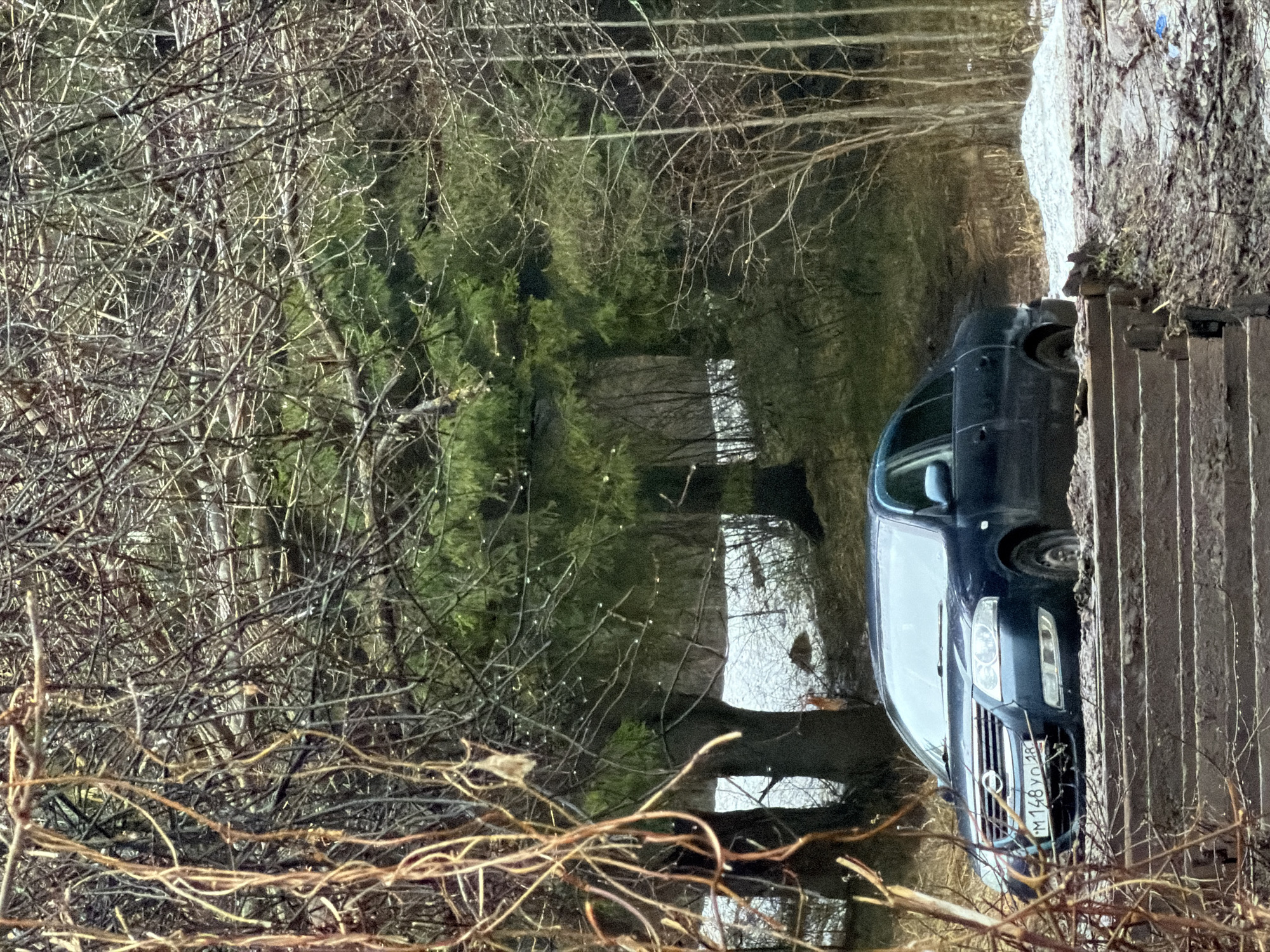 The image shows a dark blue Volkswagen Transporter van parked beside a concrete barrier, possibly a bridge or embankment. The vehicle's license plate reads "M 148 YO 18". The surroundings appear to be a natural setting, with bare trees and possibly a body of water behind the vehicle. The location is Izhevsk, Russia.