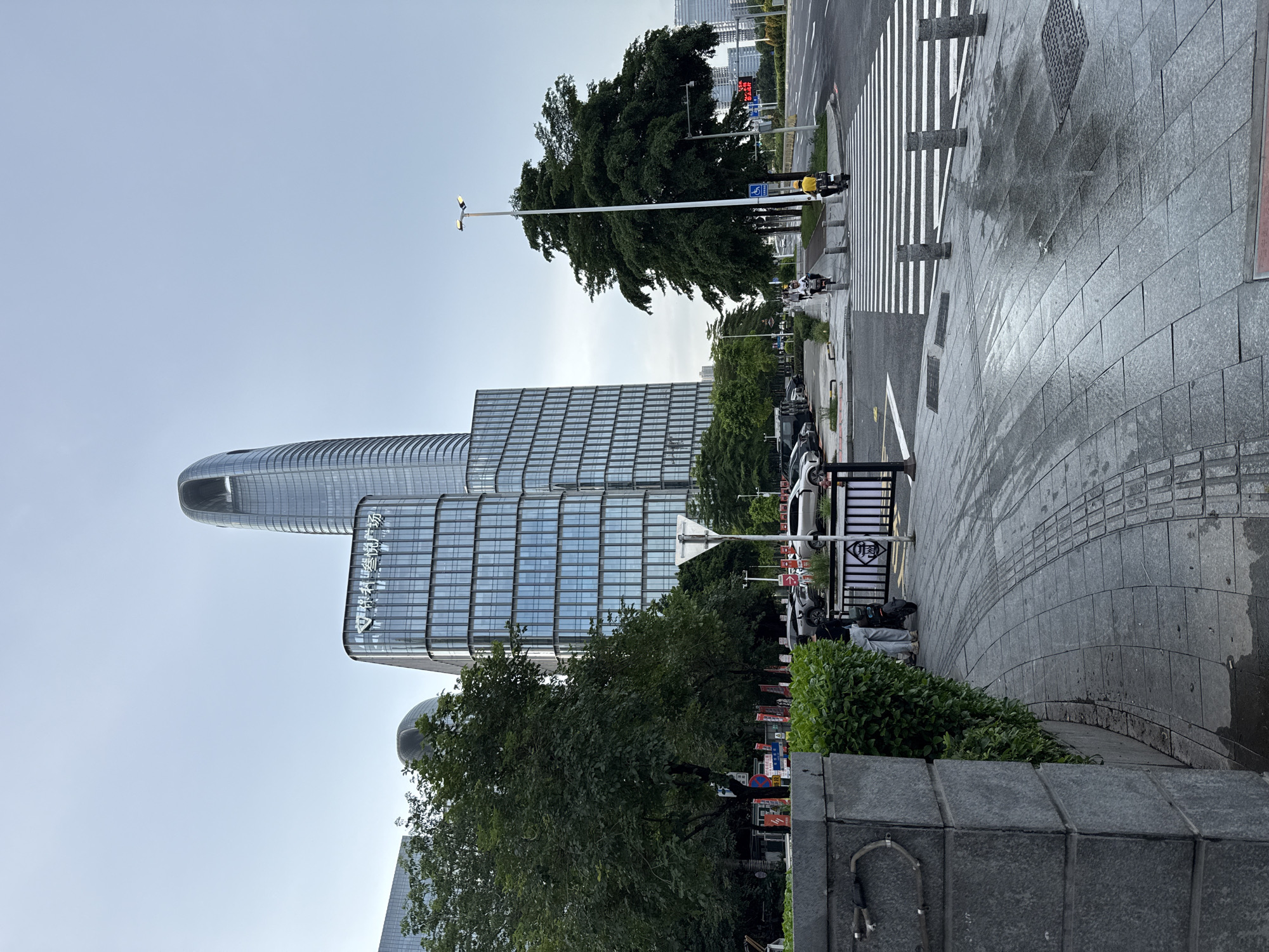 The image captures a street scene in Haizhu District, China, featuring modern architecture and urban elements. In the background, a tall, sleek skyscraper with a distinctive curved top dominates the skyline, flanked by other contemporary glass-fronted buildings. A mid-rise building with a grid-like facade and Chinese characters is also visible.
In the foreground, a wide sidewalk paved with gray stone slabs shows signs of recent rain, with visible wet patches and reflections. A stone planter with dense green foliage lines the left side of the sidewalk. Adjacent to the sidewalk, a street with multiple lanes is visible, with cars parked along the side and a pedestrian crossing marked with white stripes.
Two individuals are on the sidewalk: one person is on a bicycle, and another is on a scooter. A street lamp with two lights stands tall on the right side of the image. Several bollards are placed along the edge of the sidewalk and the pedestrian crossing. Red and white traffic signs and poles are scattered along the street. The sky is overcast and pale blue.