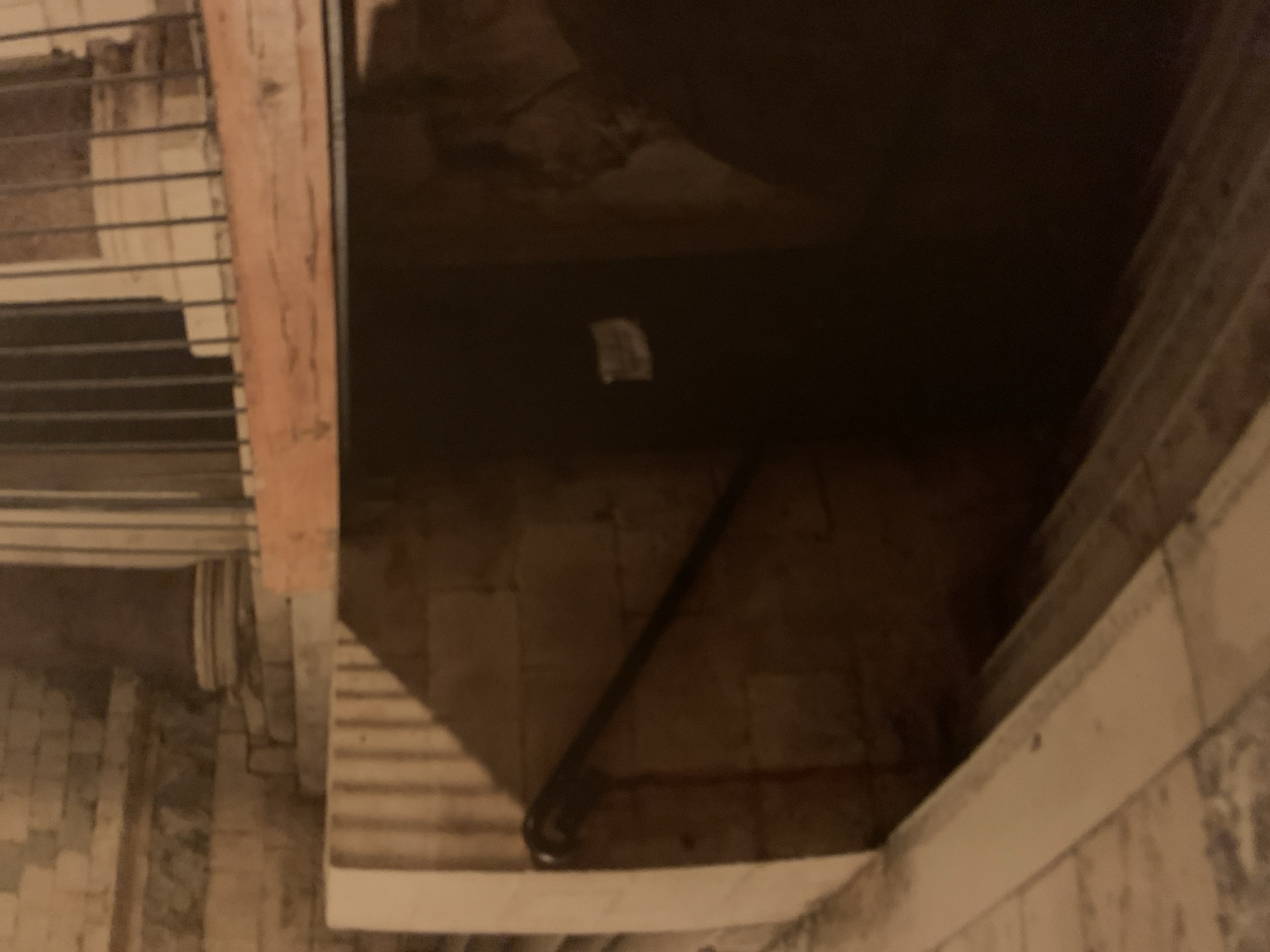 The image depicts a dark, confined space, possibly an undercroft or basement. The walls are constructed of stone or brick, with visible signs of wear and discoloration. A metal railing or pipe runs along the floor. There is a small piece of white paper on the floor as well. The location is the crypt of the Sagrada Familia church in Barcelona, Spain. A structural detail of the crypt is evident on the top left, while the lower right showcases the start of a stairway.