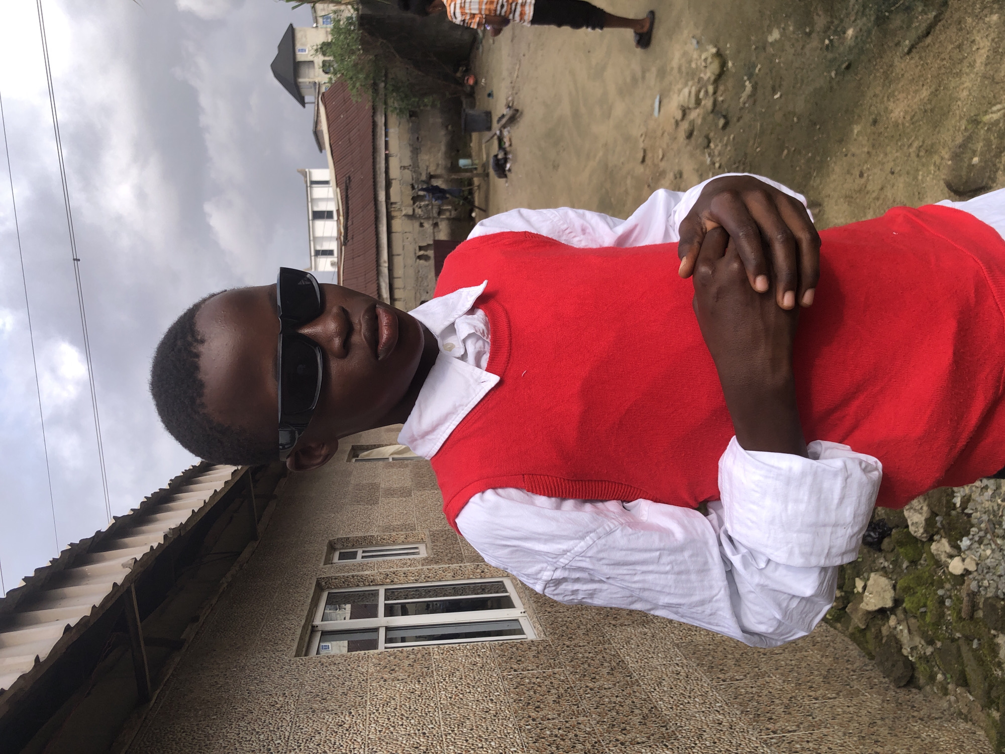 In Ado, Nigeria, a young Black boy stands before a textured, beige building with a corrugated metal roof. He wears dark sunglasses, a white button-down shirt, and a bright red sleeveless vest. His hands are clasped in front of him. The background reveals a dusty yard with scattered debris, other buildings, and a person in an orange and white striped shirt walking away. The sky is overcast with dramatic clouds.
