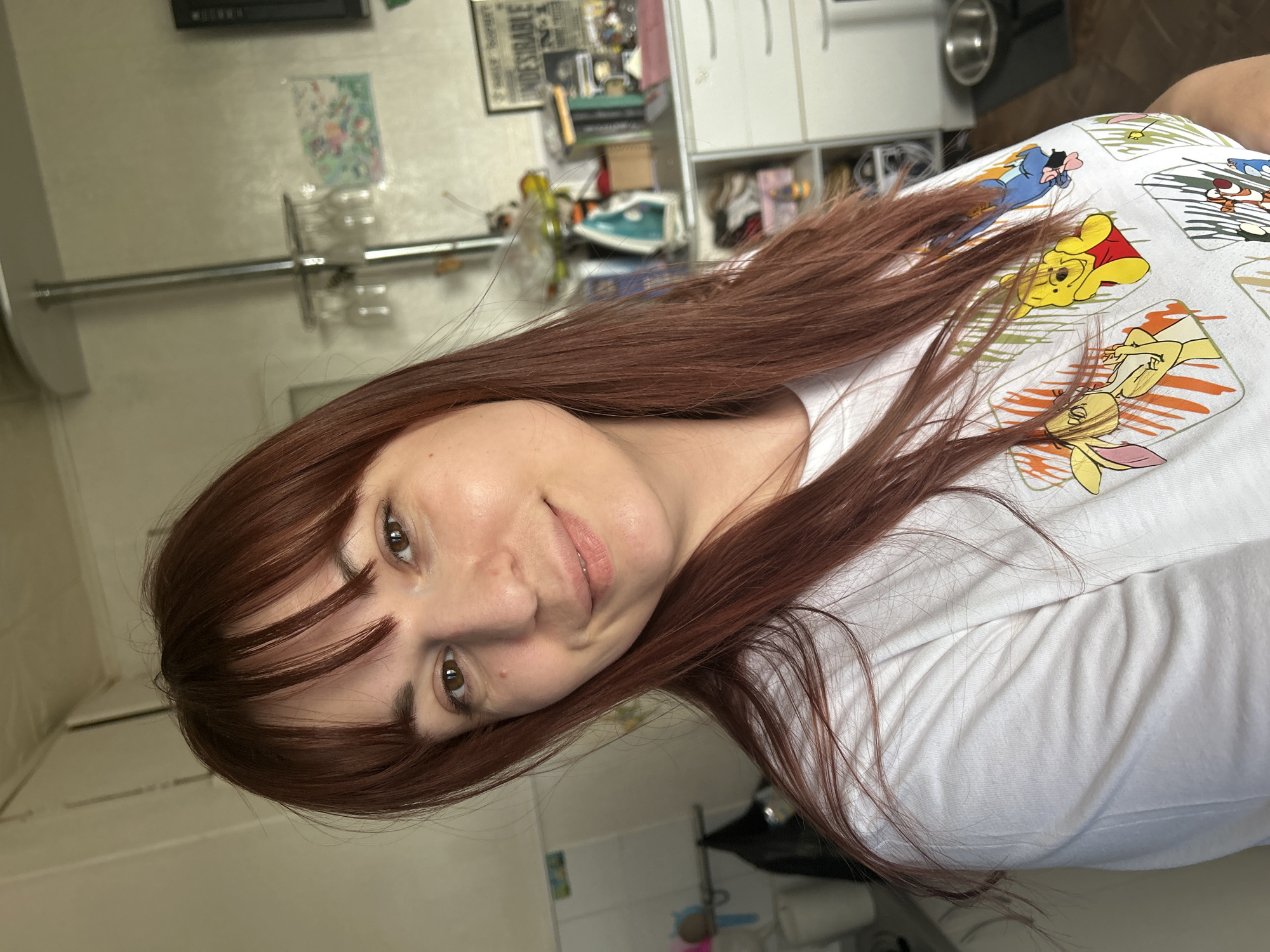 A woman with auburn hair and bangs is wearing a white T-shirt with Winnie the Pooh characters on it. The background shows a cluttered room with white cabinets, a framed print, and an iron. There is no indication of the city of Фоки, Russia, being related to the image.