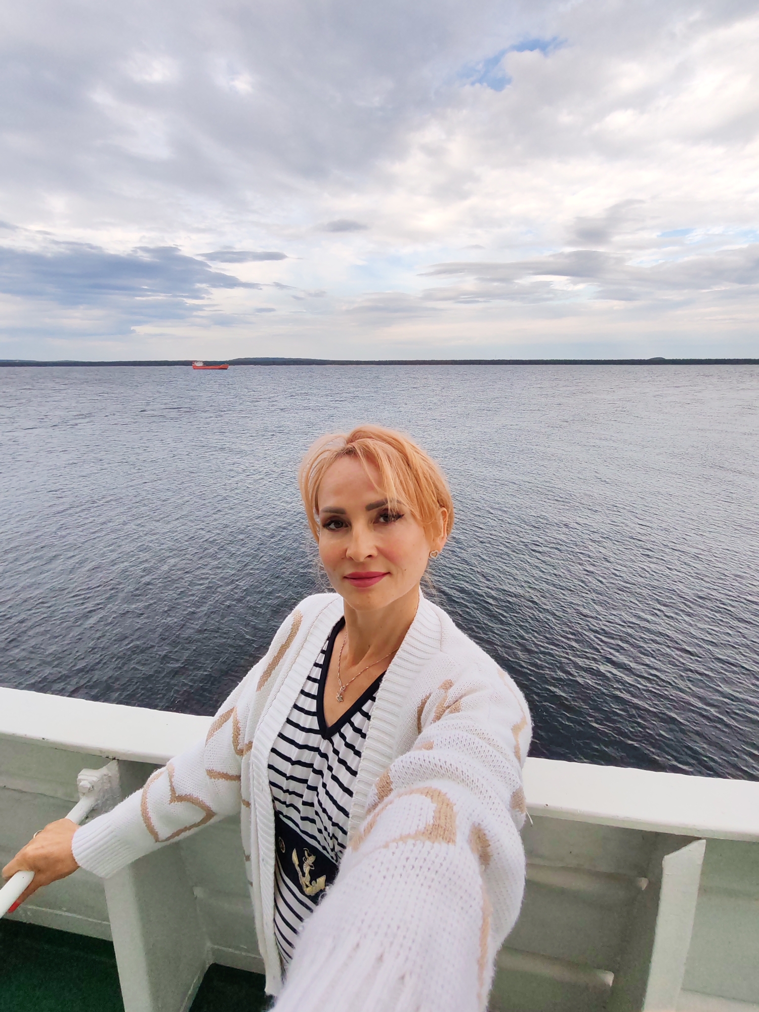 A woman with short blonde hair and light skin takes a selfie from the deck of a ship. She is wearing a white knitted cardigan with a beige geometric pattern over a black and white striped shirt. The background shows a vast body of dark water under a cloudy sky. In the distance, a small red cargo ship is visible on the horizon, and the silhouette of a landmass with trees lines the far shore. The railing of the ship is in the foreground, and a white pole is to the woman's left. The overall impression is a moment captured during a voyage on a cloudy day, with the expansive water and distant ship suggesting a journey. The location is identified as Shal'skii, Russia.