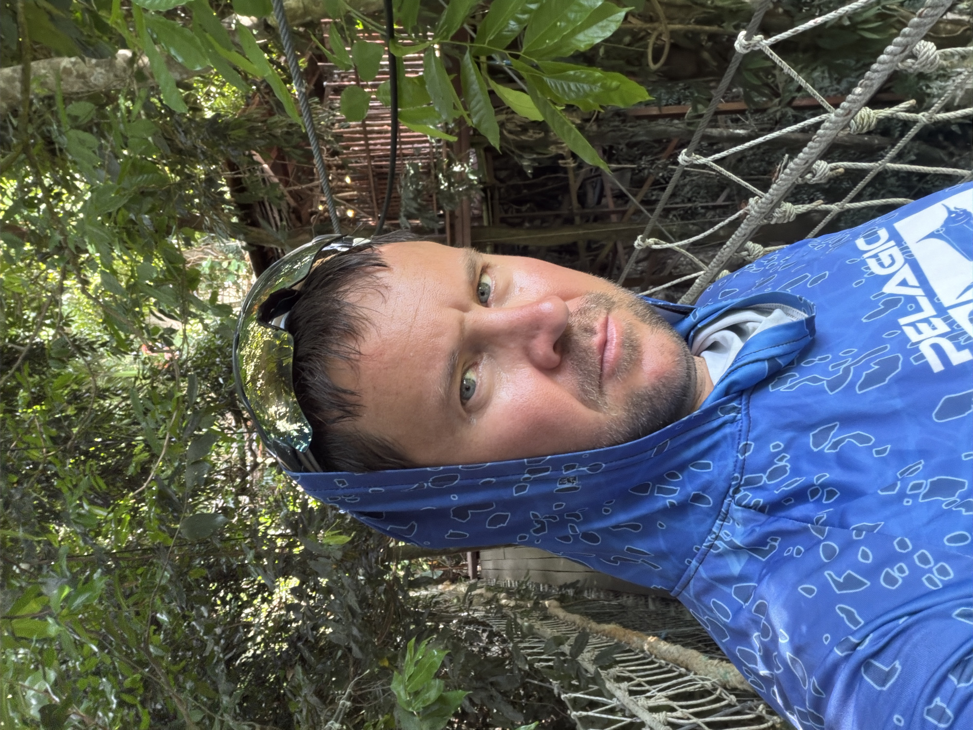 This is a portrait of a man lying down, likely in a hammock or similar suspended structure, outdoors. He is wearing a blue long-sleeved shirt with a white abstract pattern and sunglasses on his head. A blue bandana or gaiter is pulled up over his head and neck, partially obscuring his forehead. The man has light-colored eyes, stubble, and is looking towards the camera.
The background is filled with lush green foliage, suggesting a tropical or heavily wooded environment. There are also hints of man-made structures, including what appears to be a rope bridge or net-like material and some wooden or metal scaffolding in the distance, all interwoven with the natural surroundings. Sunlight filters through the leaves, creating a dappled effect. The overall impression is one of relaxation and immersion in nature. The visible text on the shirt says "PELAGIC". Given the location context of Ko Samui, Thailand, this scene likely takes place in a natural setting like a jungle or a resort with natural landscaping. The time of day appears to be daytime, with bright lighting.