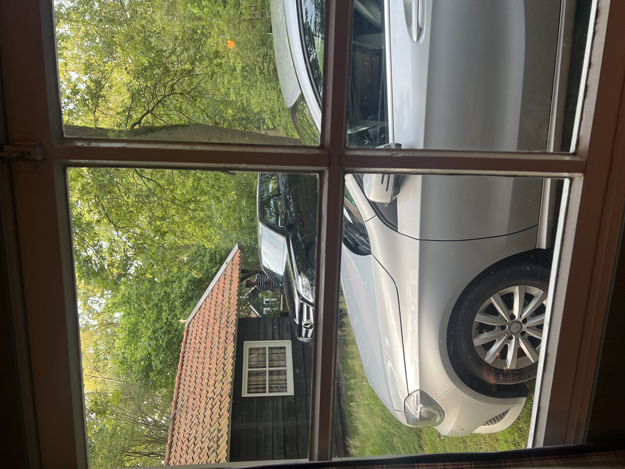 The image, taken from inside a building looking through a window with multiple panes, shows an outdoor scene in Collendoorn, Netherlands. In the foreground, the hood and wheel of a silver car are visible. Behind this, a black Mercedes-Benz SUV is parked. To the left, a dark wooden building with a terracotta tile roof and a white-framed window is present. A person wearing a striped shirt is standing near the black SUV. The background consists of lush green trees. The lighting suggests it is daytime, possibly late afternoon or early evening.