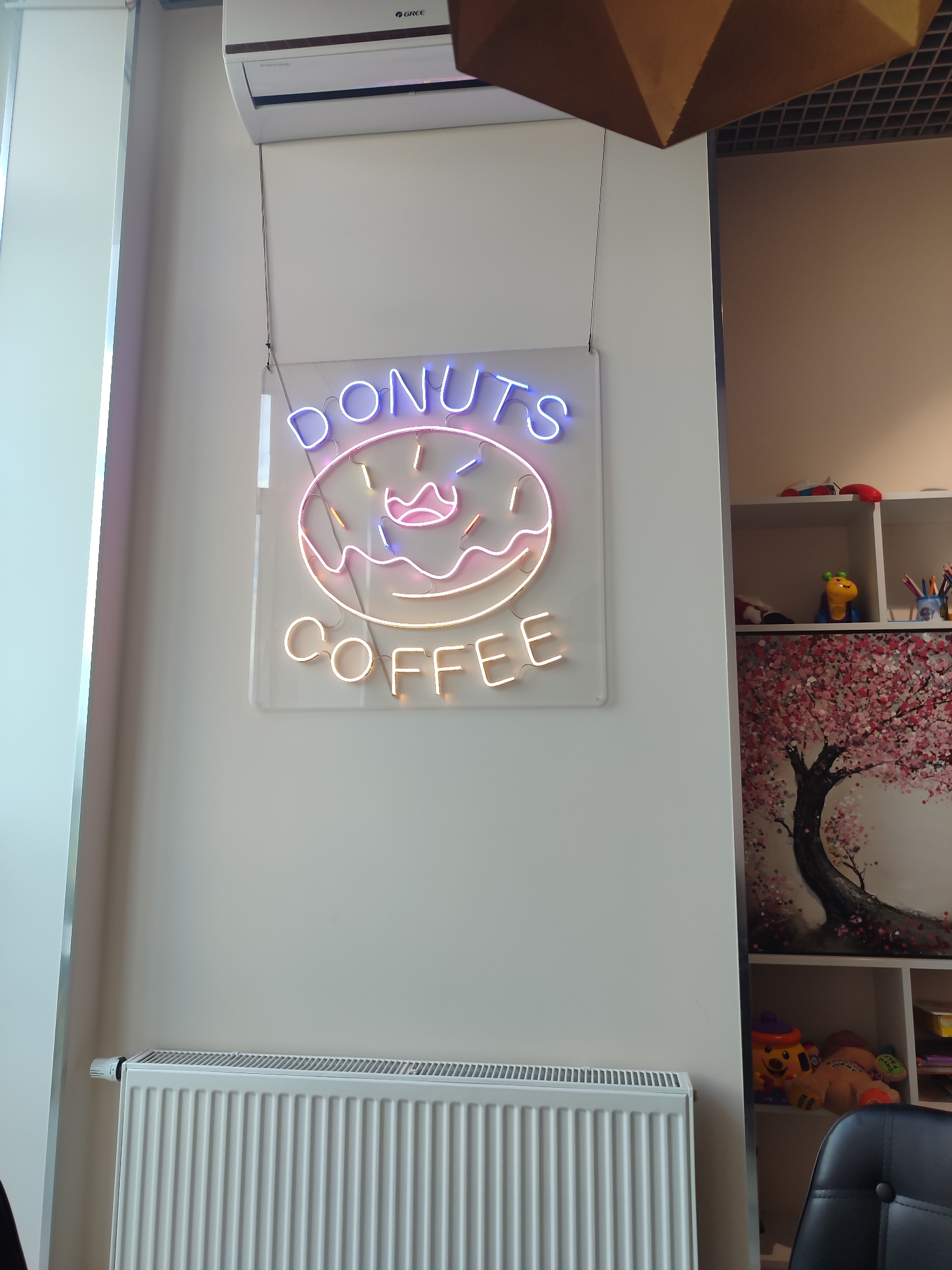 An indoor scene features a white wall with a neon sign that reads "DONUTS COFFEE" in glowing letters. The sign is shaped like a donut with icing and sprinkles. To the left of the sign, a white air conditioning unit is mounted high on the wall. Below the neon sign, a white radiator is installed along the bottom of the wall. To the right of the neon sign, a white shelving unit is visible, displaying various items including a painting of a cherry blossom tree, a yellow toy, and a stuffed animal. A black chair is partially visible in the lower right corner of the image.