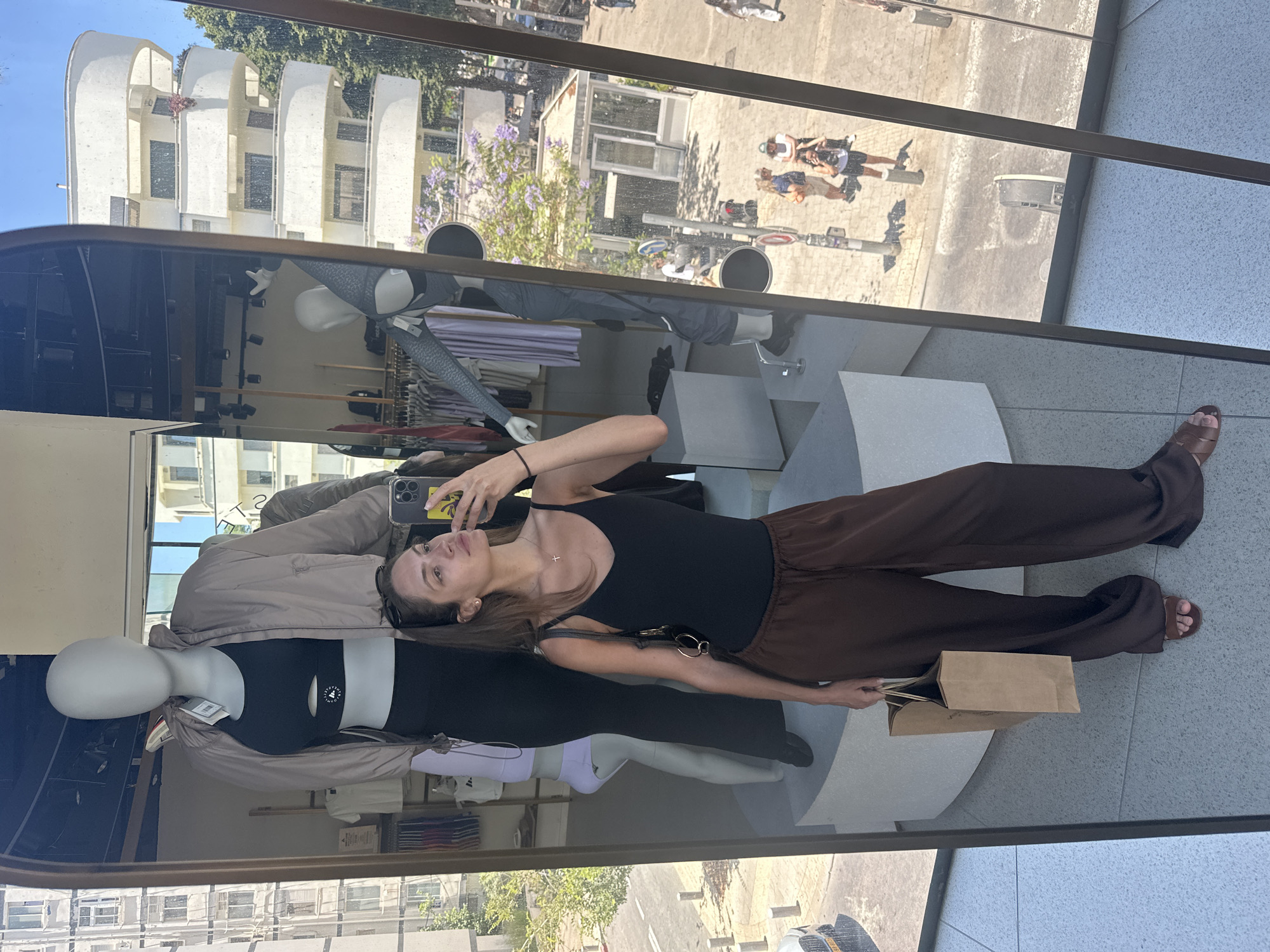 The image is a mirror selfie taken by a woman inside a retail store, with a large window reflecting and revealing an outdoor city scene.
In the foreground, a young woman with long brown hair, wearing a black spaghetti strap tank top and loose brown wide-leg trousers with sandals, holds a smartphone in her right hand to capture the reflection. She carries a brown paper shopping bag in her left hand. A small cross necklace is visible around her neck, and sunglasses rest on her head.
The interior of the store, reflected in the mirror, shows several faceless mannequins. One prominent mannequin behind the woman wears a black sports bra, black leggings, and a light brown athletic jacket with a price tag. Clothing racks with various garments are visible further inside the store. The flooring is a light-colored tile.
The exterior street scene, visible through the window and reflected in the mirror, clearly indicates the city of Tel-Aviv, Israel. Key details supporting this identification include:
1. A prominent white building with distinctive rounded balconies, characteristic of Tel-Aviv's Bauhaus architecture.
2. Trees with purple blossoms, indicative of Jacaranda trees, which are common in Tel-Aviv.
3. A car partially visible on the street displays a blue-banded license plate, consistent with Israeli vehicle registration plates. The format "72-005-24" (partially visible) further matches Israeli plate structures.
4. Pedestrians are visible on the sunny sidewalk, and a small building with the word "CONCRETE" is seen across the street.
The overall scene captures a moment of casual shopping on a bright, sunny day in an urban setting.