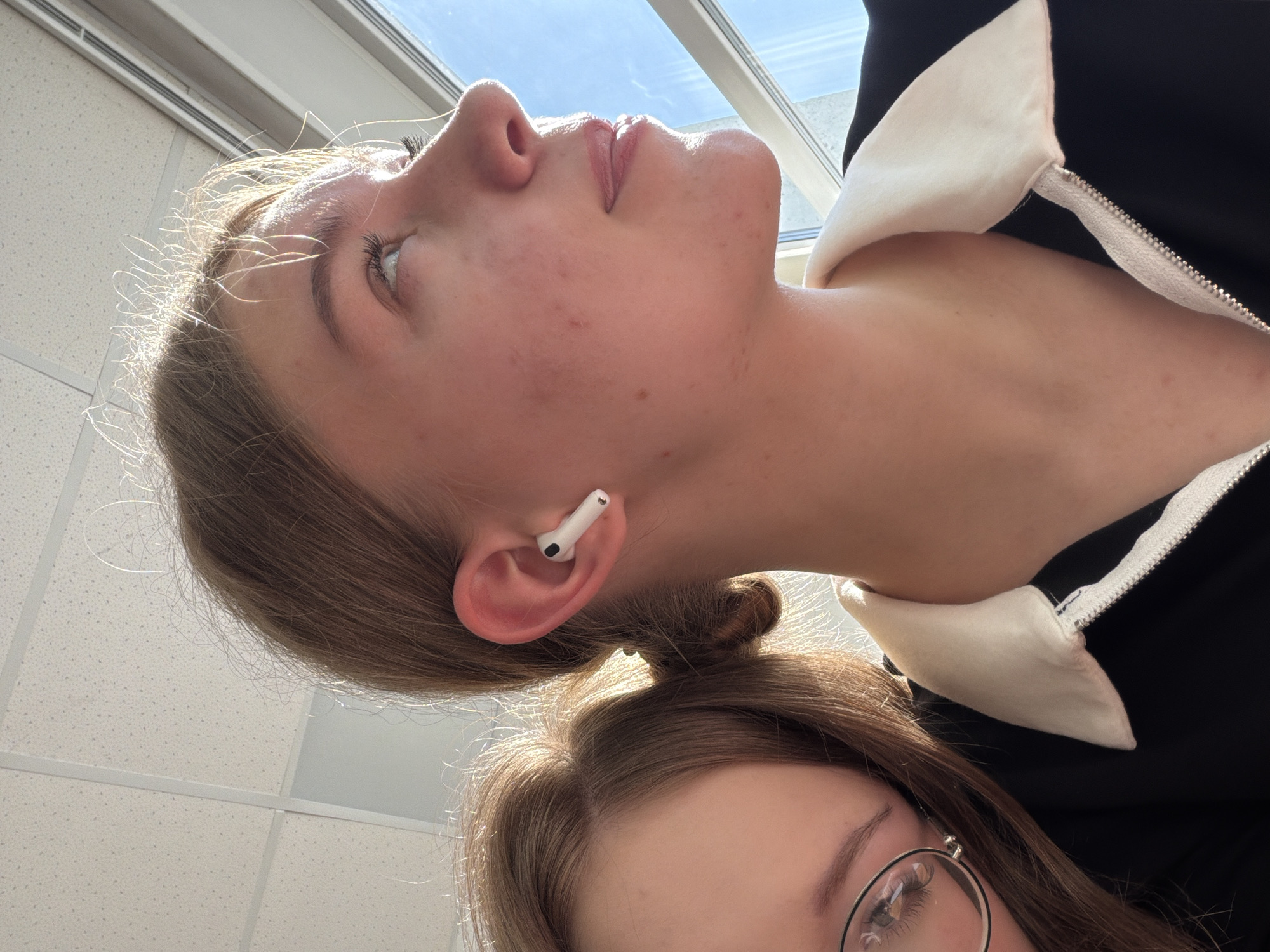 The image shows two young women in an indoor environment, likely a building in Chaikovsky, Russia, given the prompt context. One woman is in the foreground and visible from the eyes up, wearing glasses, with light brown hair. The other woman is positioned behind and above the first. Her head is tilted back, exposing her neck and a visible white earbud in her ear. She has light hair pulled back in a bun. She's wearing a black zip-up jacket with a white collar. Overhead are rectangular ceiling panels, and light is coming from a window above.