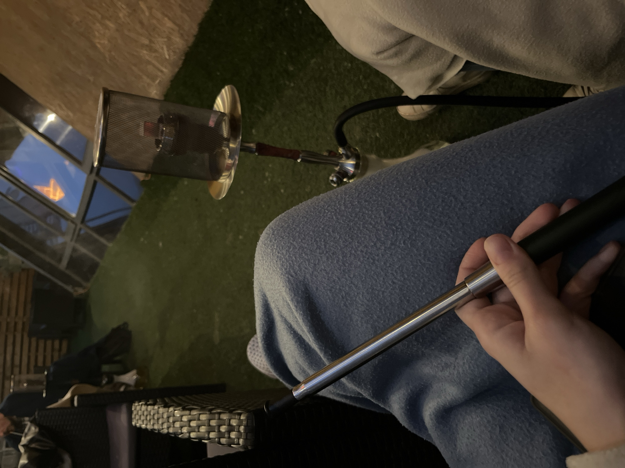 A person is holding a hookah hose in their hand, sitting on a wicker chair. The hookah is on the table next to them, with the hose connected. Another person is sitting nearby. The location is not specified.