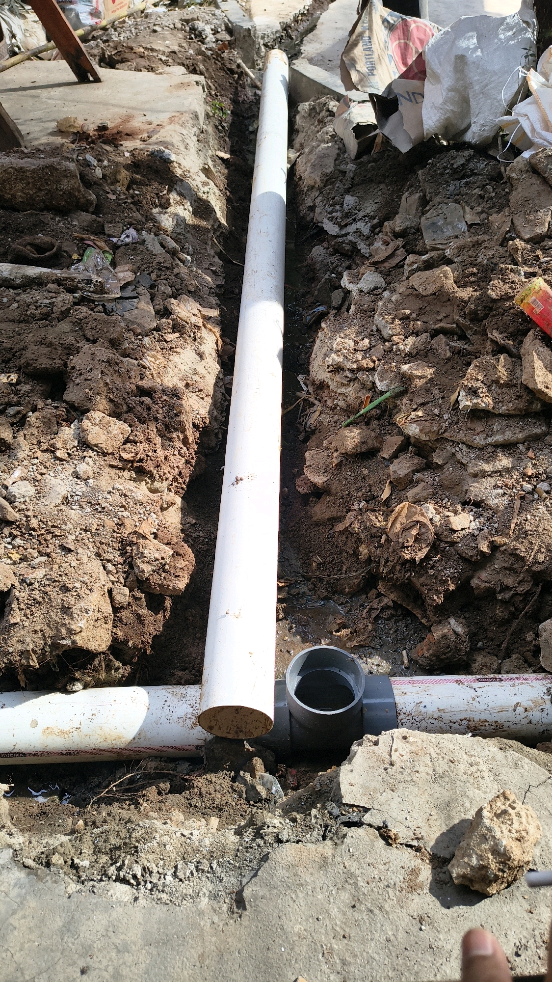 The image depicts an outdoor scene, likely a construction or repair site in South Jakarta, Indonesia. The foreground shows a partially dug trench filled with soil, rocks, and debris. Several white PVC pipes are visible, some lying horizontally and one vertically. A grey PVC fitting, possibly a T-junction or a connector, is also present in the trench. The background reveals more of the trench, with piles of dirt and scattered construction materials, including bags of cement. The lighting suggests it is daytime. A hand is partially visible in the bottom right corner of the image, indicating human presence at the site.