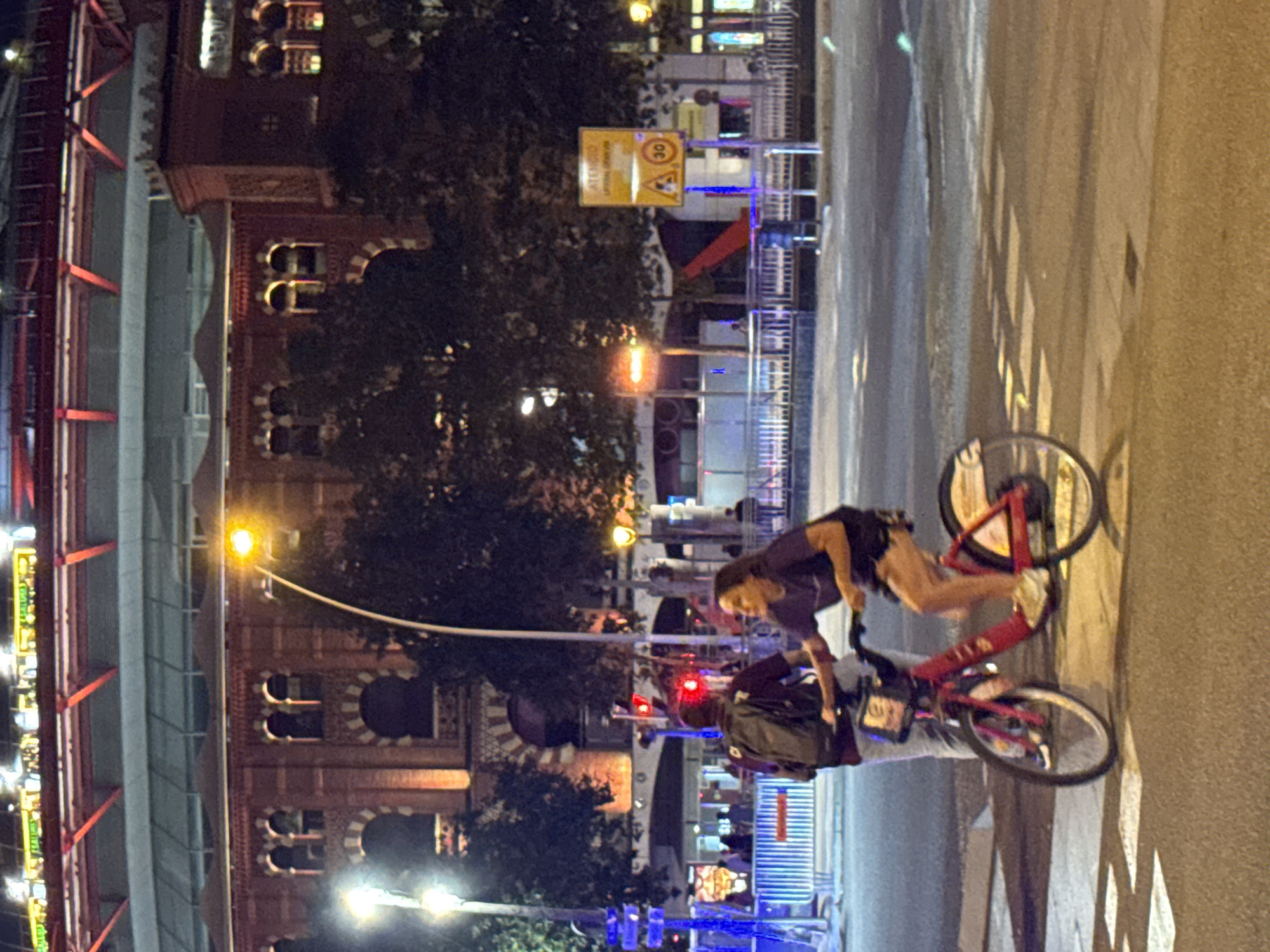 The image captures a street scene at night in Barcelona, Spain, likely near an arena or large public venue, given the architectural style of the building in the background which resembles bullfighting arenas often found in Spain. In the foreground, a woman is riding a red public-use bicycle with a basket. She is wearing a dark t-shirt and shorts. Behind her, a person wearing a dark t-shirt and a backpack is standing, holding onto the back of the bicycle. They are positioned on a crosswalk. The background shows a large, brick building with arched windows, illuminated by lights. Streetlights are visible, casting a warm glow, and trees with dark foliage are present. A yellow road sign with a red circle and the number "30" is visible on the right side of the image, indicating a speed limit. Red and blue lights from traffic signals or temporary barriers are also visible. The overall impression is a lively urban setting at night, with people navigating the streets on a shared bicycle system.