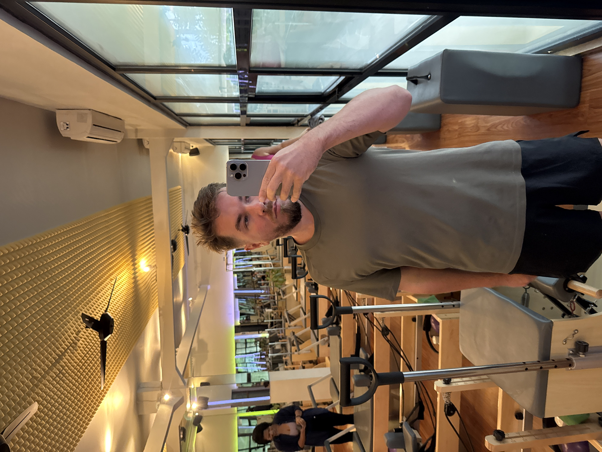 A man is taking a mirror selfie inside what appears to be a modern Pilates or fitness studio. He is standing upright, holding a grey smartphone with multiple camera lenses to capture his reflection in a large wall mirror. He is wearing a dark olive-green t-shirt and dark shorts, and has a beard.
The studio environment, as seen in the reflection, features several Pilates reformers with light wooden frames and grey padded surfaces, arranged in rows. A prominent glass panel or skylight structure forms part of the ceiling, allowing natural light into the space. An air conditioning unit is mounted on a white beam near the ceiling. On a side wall, a section is covered with golden, textured acoustic paneling, where a minimalist wall clock with dark hands is mounted. Another air conditioning unit is visible further back. The floor is made of warm-toned wood.
In the background reflection, another person, a woman with dark hair, is visible. She appears to be interacting with one of the Pilates reformers or possibly looking at her phone. She is wearing dark trousers and a light-colored top under an open dark shirt. The overall atmosphere of the studio is bright and clean, with a mix of natural and artificial lighting. No discernible text is visible within the image. The location context provided is North Kuta, Indonesia.
