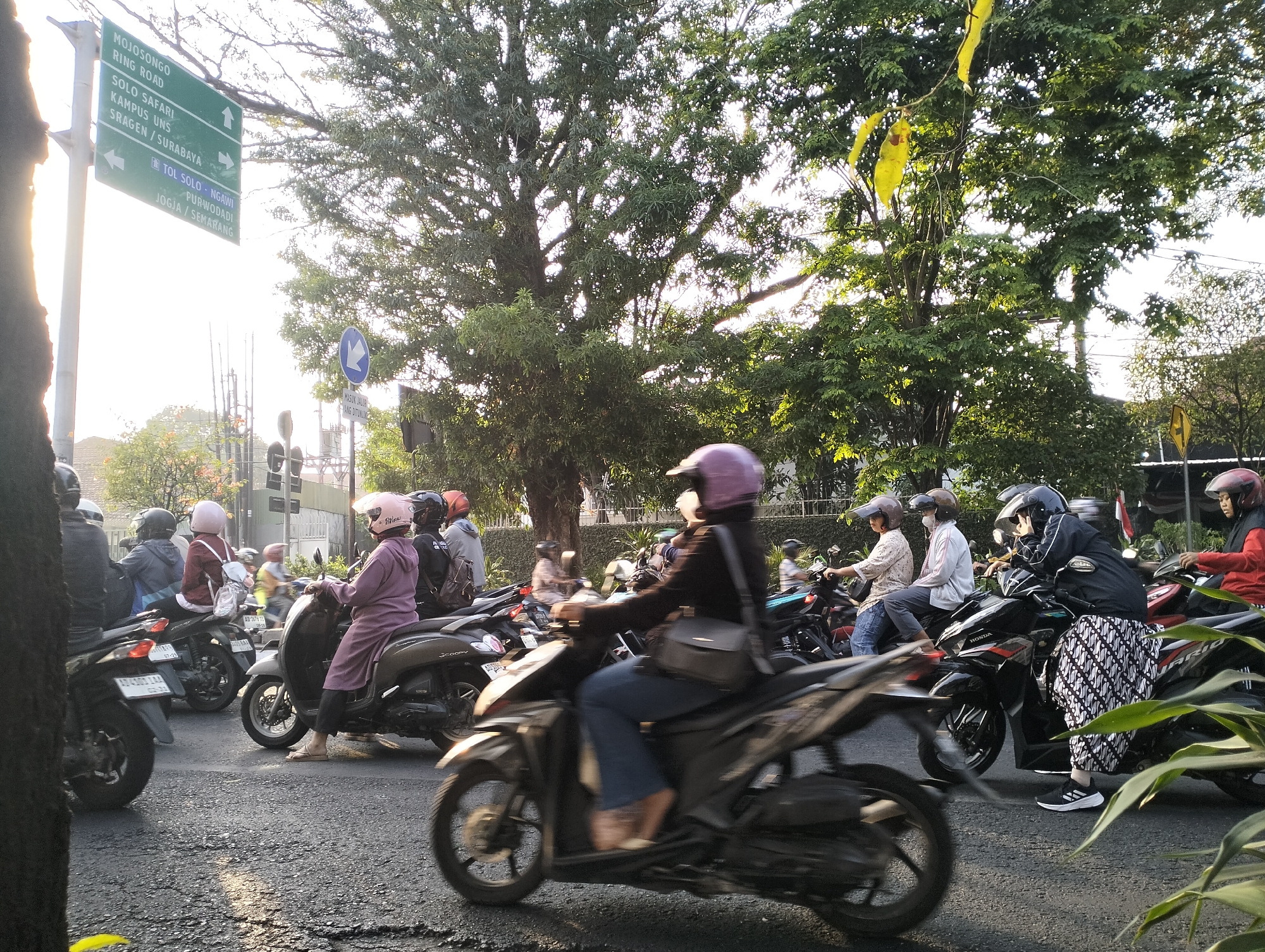 In Surakarta, Indonesia, a group of motorcyclists navigate a street with lush trees and signage. The motorcyclists, most wearing helmets, are seen on various models of scooters and motorcycles. One person in a purple tunic rides a scooter, while another in a maroon jacket and pink helmet is on the back of a motorcycle. Traffic is moving, indicated by the blur of motion on the vehicles and people. A prominent green street sign points towards "MOJOSONGO RING ROAD," "SOLO SAFARI," "KAMPUS UNS," and "SRAGEN/SURABAYA," with further directions to "TOL SOLO-NGAWI," "PURWODADI," and "JOGJA/SEMARANG." The environment suggests a moderate urban setting with abundant greenery and the presence of utility poles and wires. The lighting indicates it is likely late afternoon or early morning with the sun casting a warm glow.