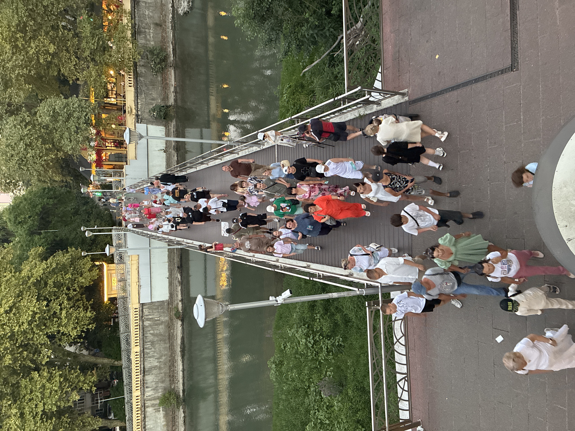 An aerial, high-angle, full shot captures a bustling pedestrian bridge over a river in Sochi, Russia, at dusk. Numerous people, in casual summer attire, traverse the bridge in both directions. The bridge is lined with metal railings and streetlights, and a camera is mounted on one of the lampposts. Lush green trees and vegetation flank the riverbanks, with buildings and illuminated businesses visible in the background. The scene suggests a lively, public gathering space.