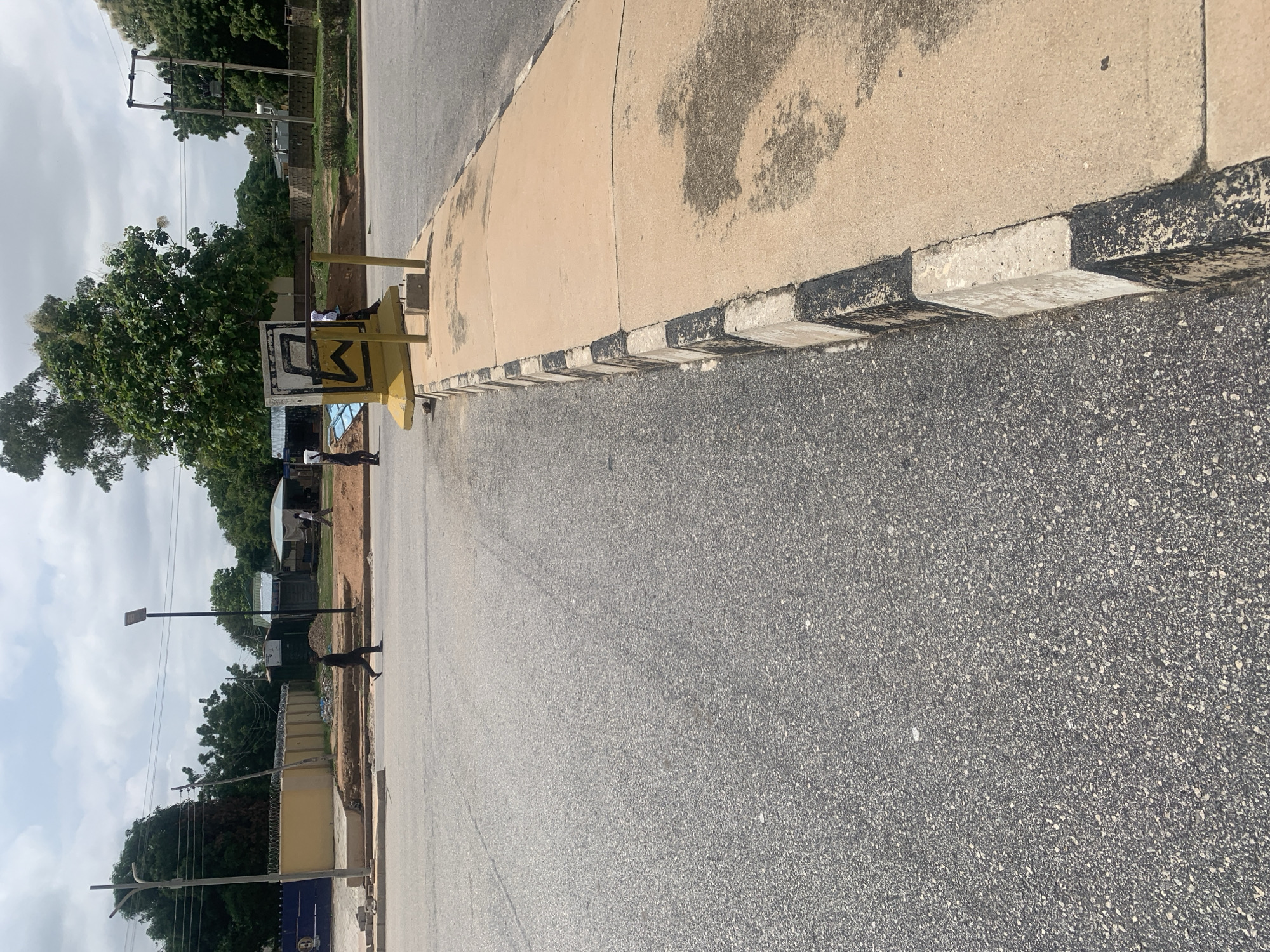 The image depicts a street scene in Jalingo, Nigeria. In the foreground, a paved road runs diagonally from the lower left to the upper right. To the right of the road is a raised sidewalk with a concrete curb that is painted with alternating black and white stripes. A yellow sign with black geometric markings stands on the sidewalk, supported by metal poles.
Further in the background, several individuals are visible walking. One person is crossing the road, and two other figures are on the sidewalk near the sign. Beyond the sidewalk, there are buildings with corrugated metal roofs, some construction materials, and trees. A blue fence with barbed wire runs along a property line. Utility poles and power lines are also present, along with a street light. The sky is overcast.