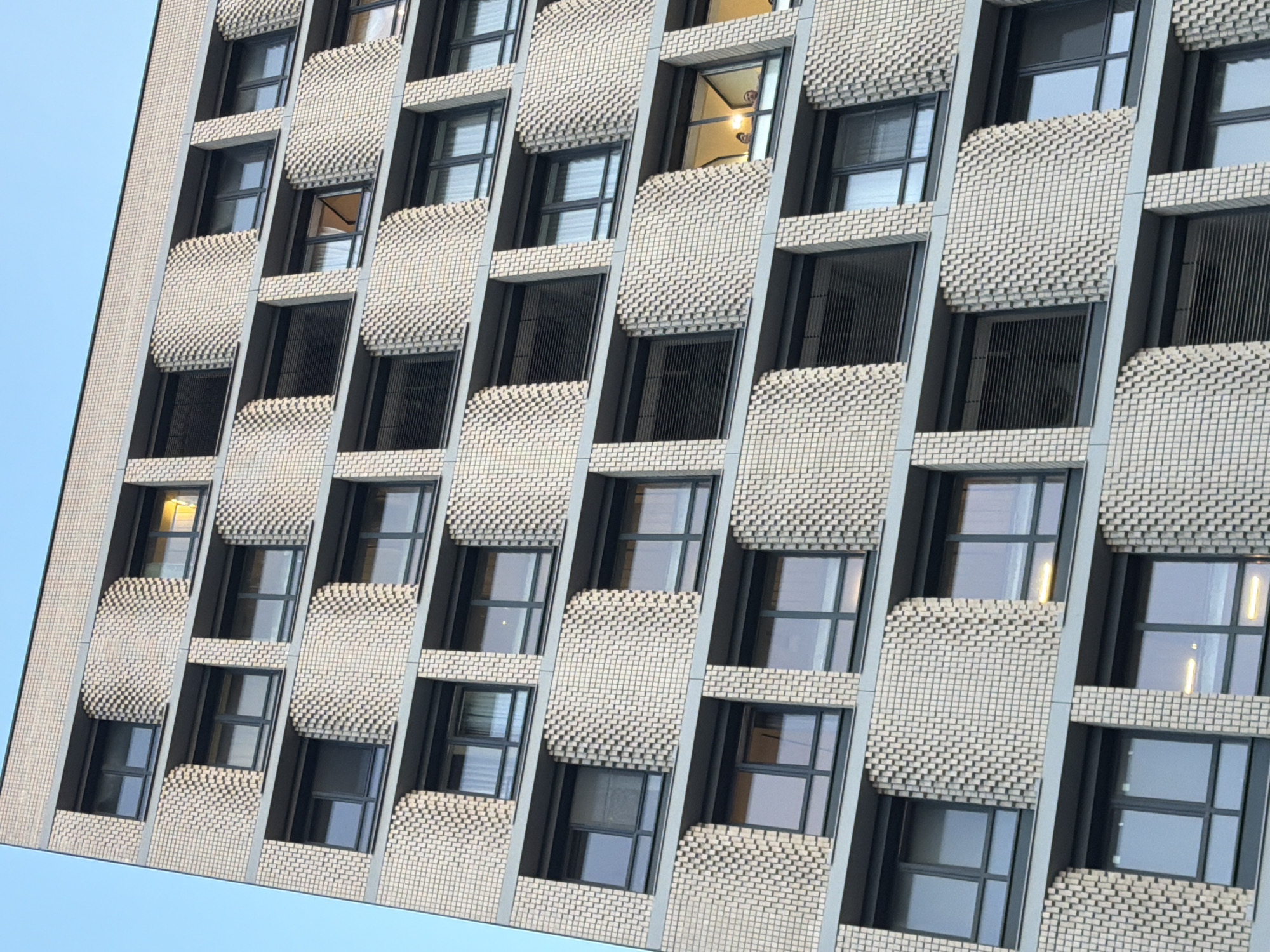 A low-angle, full-frame photograph captures the exterior of a modern apartment building located in Moscow, Russia. The building features a repetitive pattern of light-colored brickwork with recessed windows. The facade is broken by textured, curved brick elements that alternate with flat window panels. The windows are dark-framed and vary in transparency, with some showing interior blinds and others revealing glimpses of illuminated interiors. In one of the upper windows, two individuals are visible, looking out. The sky above is a clear, pale blue.