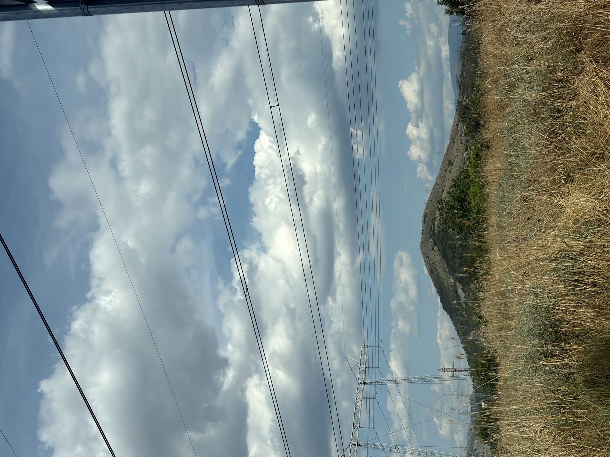 An eye-level, medium shot captures a landscape dominated by power lines and a grassy foreground. In the foreground, dry, golden grass and a few patches of green foliage fill the lower half of the frame. Beyond the grass, a gently sloping, barren hill with sparse vegetation rises in the mid-ground, its peak rounded. Two large, white electrical pylons stand to the left of the hill, their lattice structures visible against the sky. Several dark power lines stretch horizontally across the upper portion of the image, obscuring parts of the sky. The sky itself is a mix of bright blue and large, white, fluffy clouds, with some darker, gray clouds also present. In the background, more hills are visible, stretching towards the horizon. A single, dark vertical pole or antenna is visible on the far right edge of the image. The scene appears to be outdoors in a rural or semi-rural setting under a partly cloudy sky.