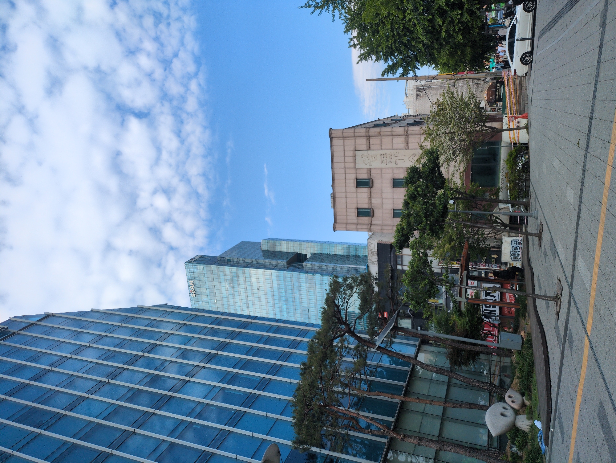 The image depicts an urban street scene in Seoul, South Korea, captured on a clear day with blue skies and scattered clouds. Two primary buildings dominate the view: a modern glass skyscraper with horizontal lines and a partially visible "SFAH" logo, and a shorter, older building with smaller windows and Korean characters on its facade.
A sidewalk runs along the right side of the frame, paved with grey stones and a yellow line. Several trees and bushes line the street, partially obscuring the view of the buildings. Signs with Korean text are visible along the street, and a white car is parked further down the street.
No people are prominently featured in the foreground. The overall impression is of a typical urban environment in Seoul.