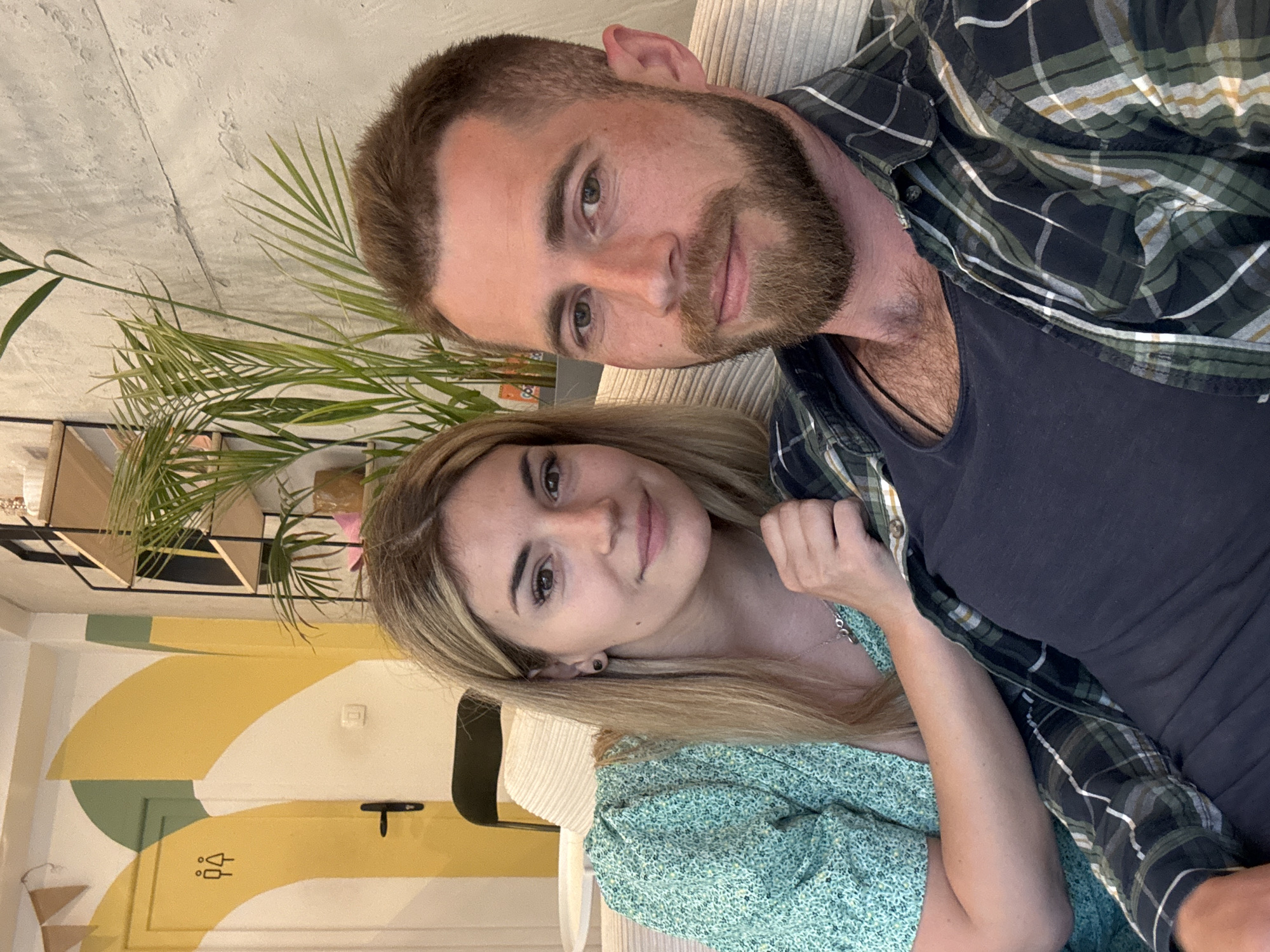 A man and a woman are posing for a selfie indoors. The man has short brown hair and a beard, wearing a dark blue tank top and a green and black plaid shirt. The woman has long blonde hair and is wearing a green floral dress. The background includes a concrete wall, a plant, and a painted door with a restroom symbol. Based on the style and decor, the location is likely a cafe or restaurant in an urban setting, potentially in Budva, Montenegro. No other specific information about the location can be determined from the image.