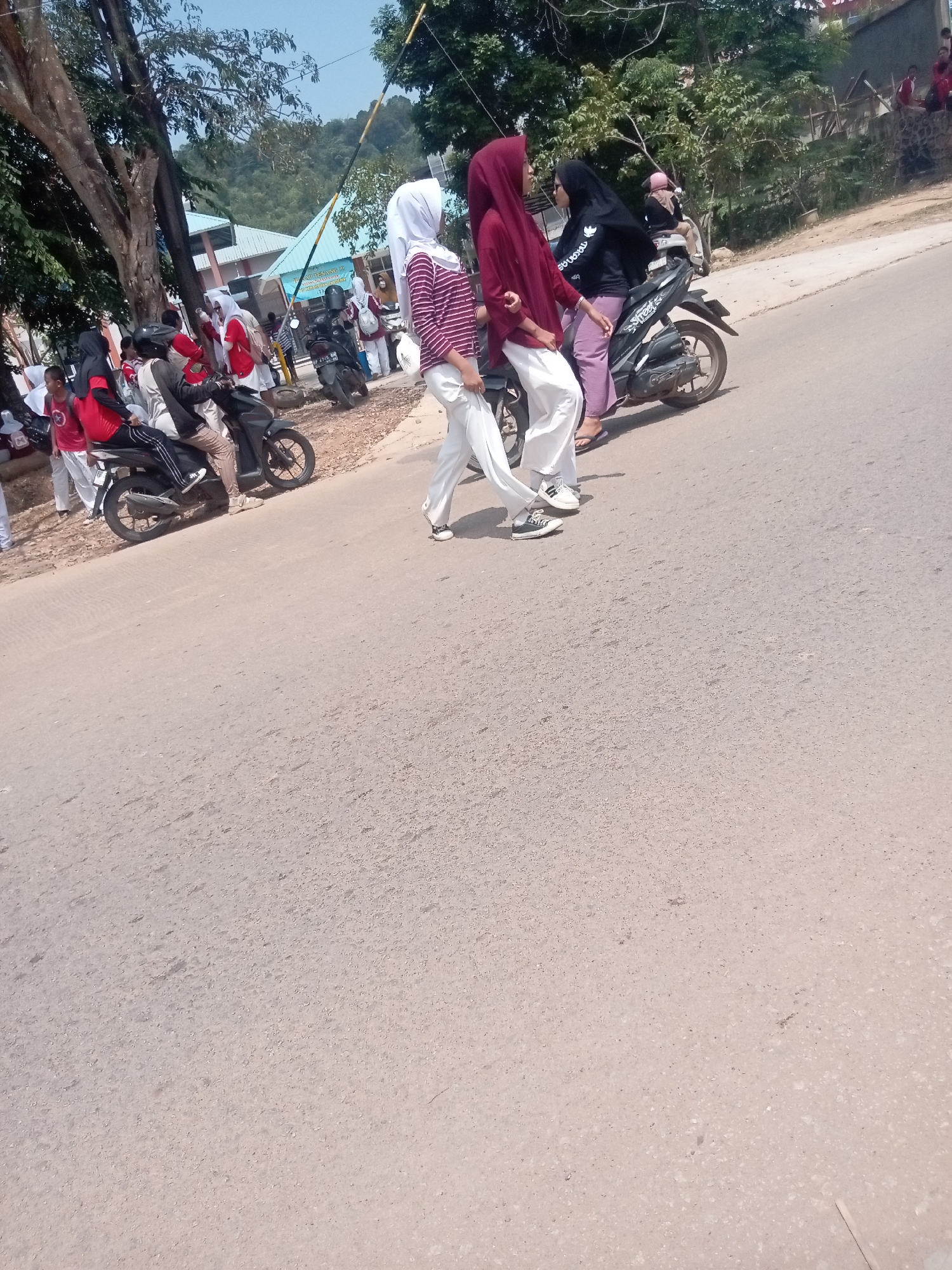 In Batam, Indonesia, on a paved road with asphalt and gravel on the shoulder, several individuals are gathered, some on or near motorcycles. A woman in a maroon hijab and a striped long-sleeved shirt, paired with white pants and sneakers, walks across the road. To her right, another woman in a black hijab and shirt, with pink pants and sandals, rides a black motorcycle. Behind them, a third person in a maroon hijab and pink pants rides a motorcycle with a passenger. Several other people, some in red shirts, are visible on the left side of the road, near parked motorcycles. Trees line the side of the road, and a building with a blue roof is visible in the background, suggesting a school or public institution. The sky is clear and blue, indicating fair weather.