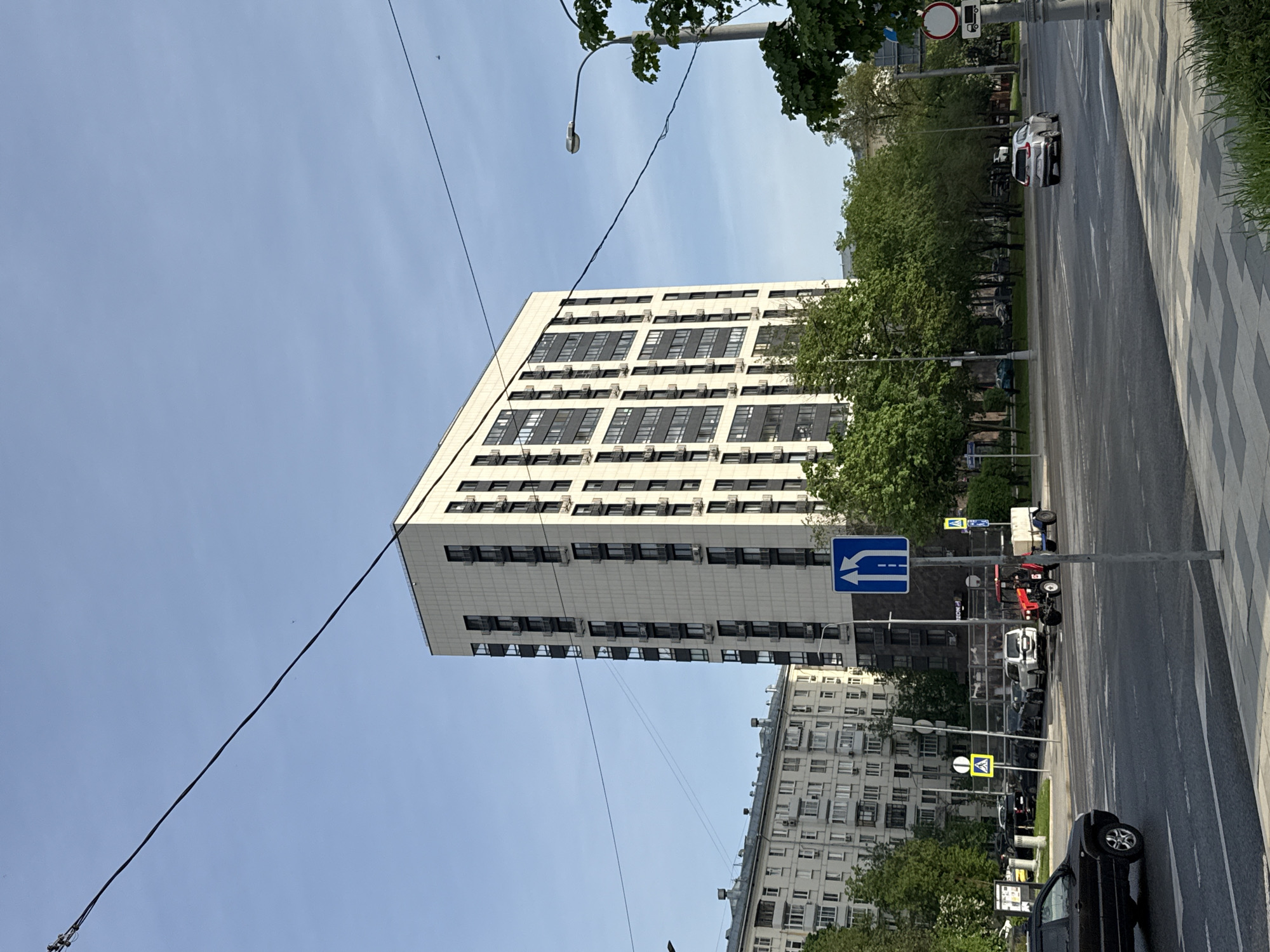 The image depicts a street scene in Moscow, Russia. A modern high-rise building with white facade and dark windows dominates the left side of the frame, ascending diagonally. Air conditioning units are visible under many of the windows. An older, multi-story residential building with smaller windows is situated to the right and slightly behind the modern building. Utility lines stretch across the sky.
On the right side of the image, a street with pedestrian zone runs along. Cars are parked along the side of the road and traffic direction sign is visible next to the pole. A small red tractor is also parked on the side of the road. Trees line the street. The sky is clear and blue.