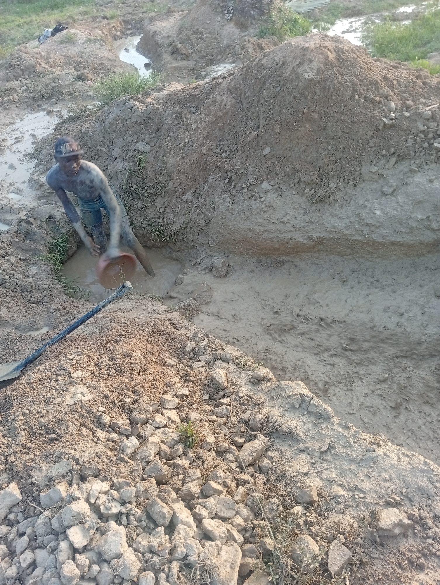 A person covered in mud is digging in a muddy trench, using a bucket to scoop out water. The scene appears to be an outdoor construction or excavation site, possibly in the city of Jos, Nigeria, given the earthy tones and the context of manual labor often associated with such environments. The ground is uneven, with piles of dirt and rocks surrounding the trench. Vegetation is sparse, with some dry grass visible. The overall impression is one of hard physical work in a challenging, wet environment.