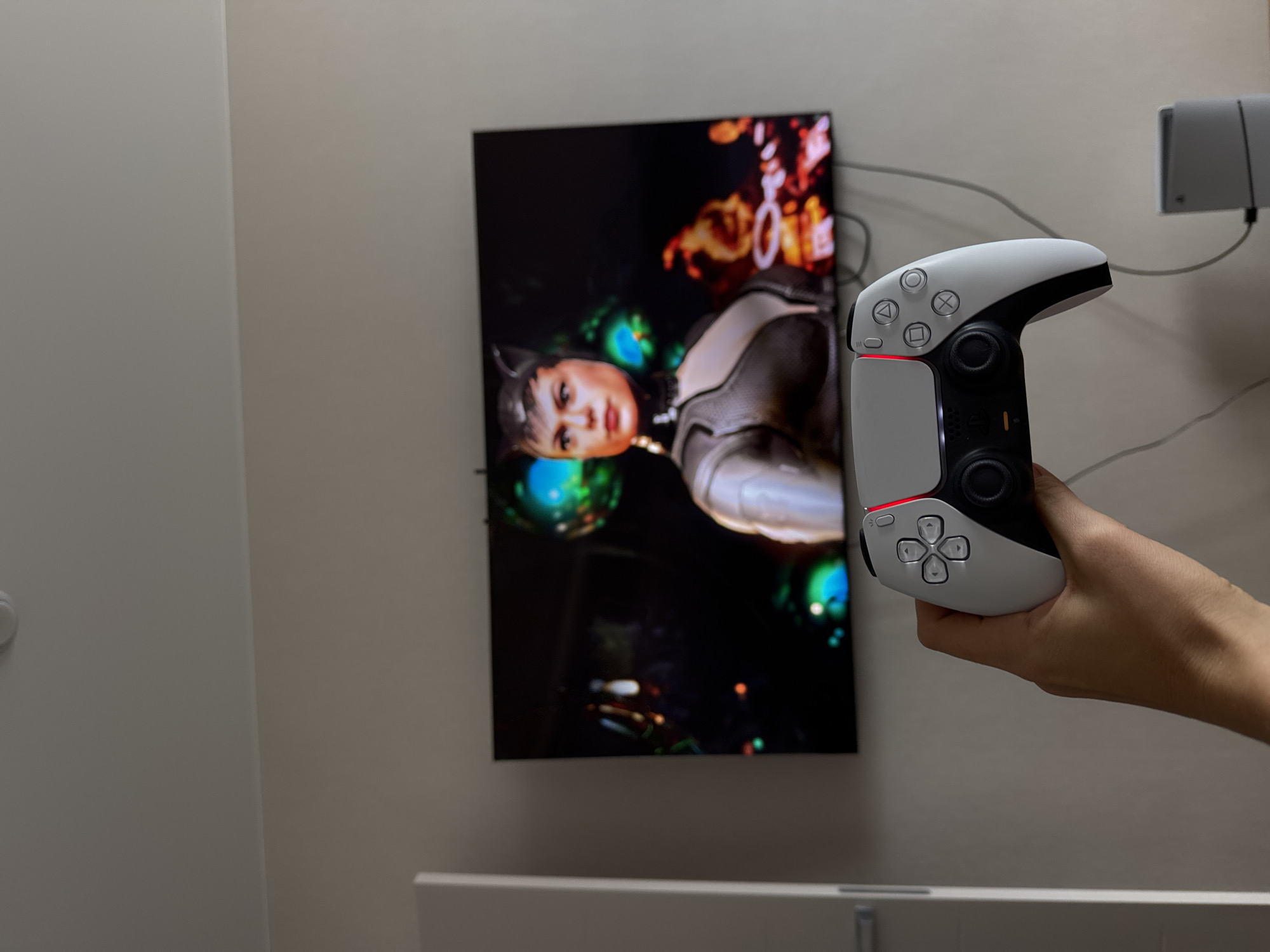 A person is holding a PlayStation 5 controller in front of a television. The television is displaying an image of a woman in a black and silver suit. There is a wall-mounted electronic device, possibly a game console, on the right side of the frame. The setting appears to be indoors, possibly a living room or entertainment space. The location is unknown.