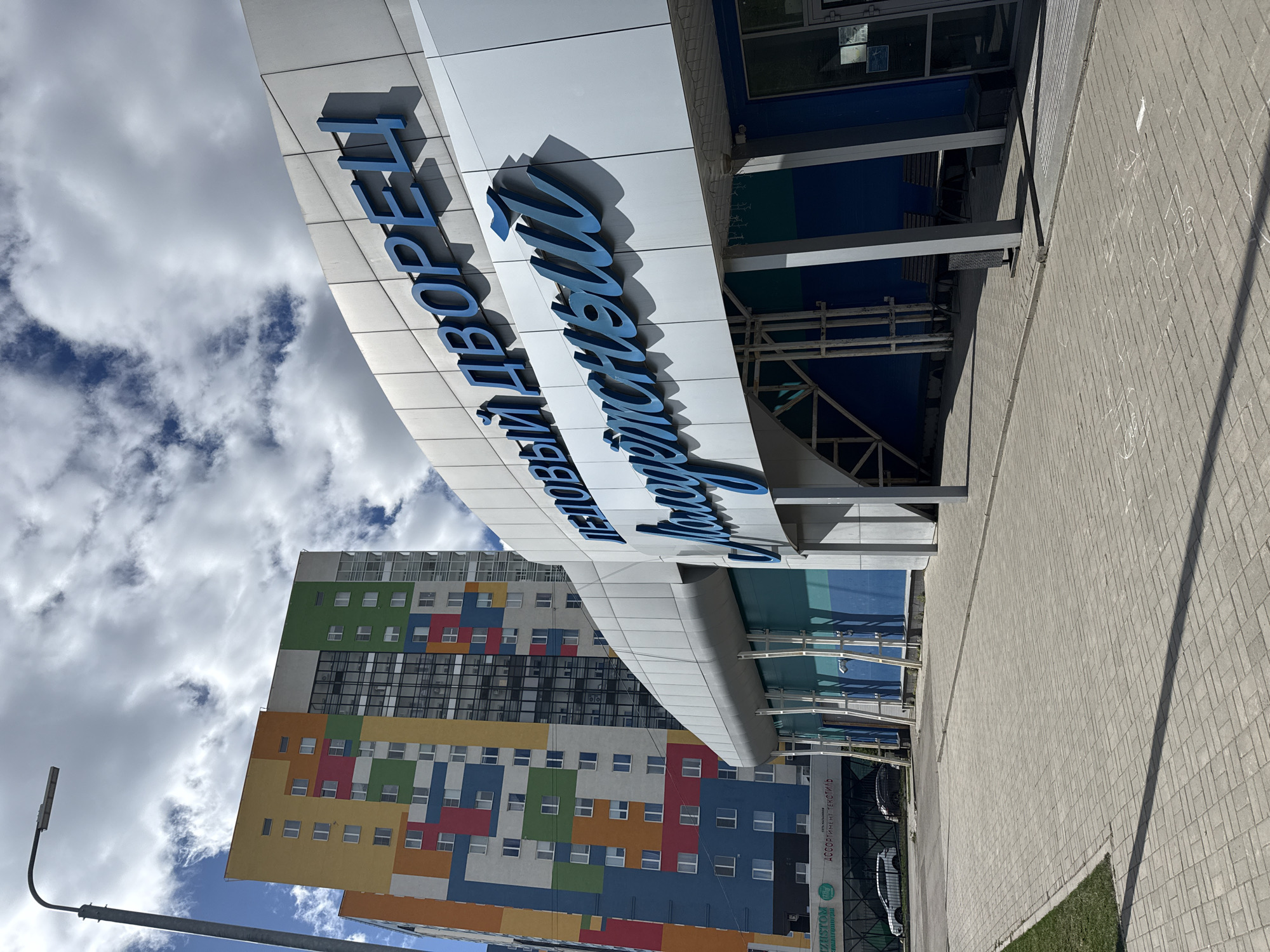 The image shows a building in Izhevsk, Russia. The facade of the building features blue lettering that reads "ЛЕГОВЫЙ ДВОРЕЦ ЧЕМПИОН". Behind the building is a colorful high-rise residential building with rectangular blocks of yellow, blue, red, green, and gray. A sidewalk is visible in the foreground. There are light clouds in the sky.