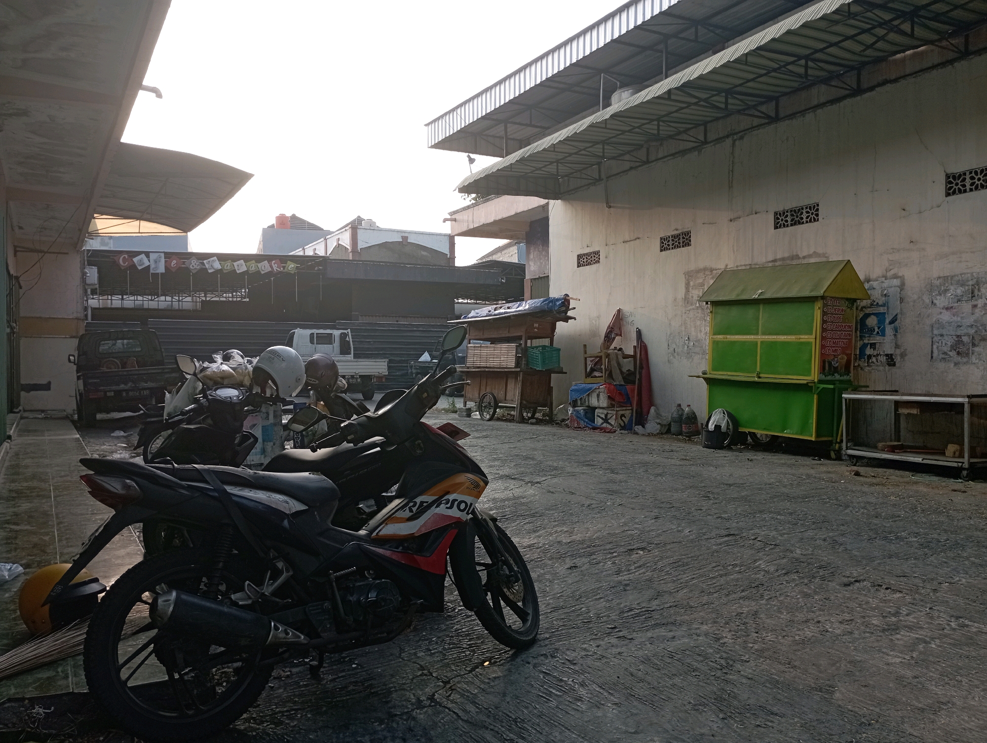 The image depicts a street scene in what appears to be a market or commercial area. In the foreground, a black motorcycle with red and orange decals is parked on a textured asphalt surface. To the left of the motorcycle, a yellow helmet and a broom lie on the ground. Behind this motorcycle, several other motorcycles, some with helmets on them, are parked. A black pickup truck is visible further back, to the left.
The background features buildings with various roof structures, including corrugated metal and tiled roofs. A prominent green food stall with yellow trim is situated against a weathered, off-white wall with several circular ventilation grates. Posters and papers are affixed to this wall. To the right of the green stall, a metal-framed display case or cart is present. The overall environment suggests an outdoor commercial space, potentially in Indonesia, given the context of Ponorogo. The lighting indicates it might be early morning or late afternoon due to the soft, directional sunlight. No people are clearly visible in the image, though the presence of parked vehicles and the food stall suggests commercial activity.